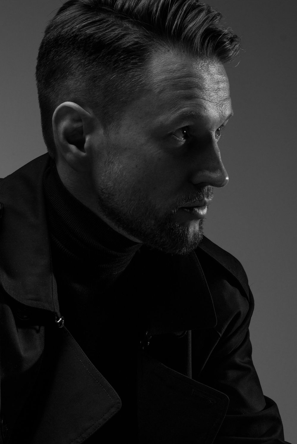 A dramatic black and white profile portrait in moody lighting features a silhouetted subject wearing a dark jacket.