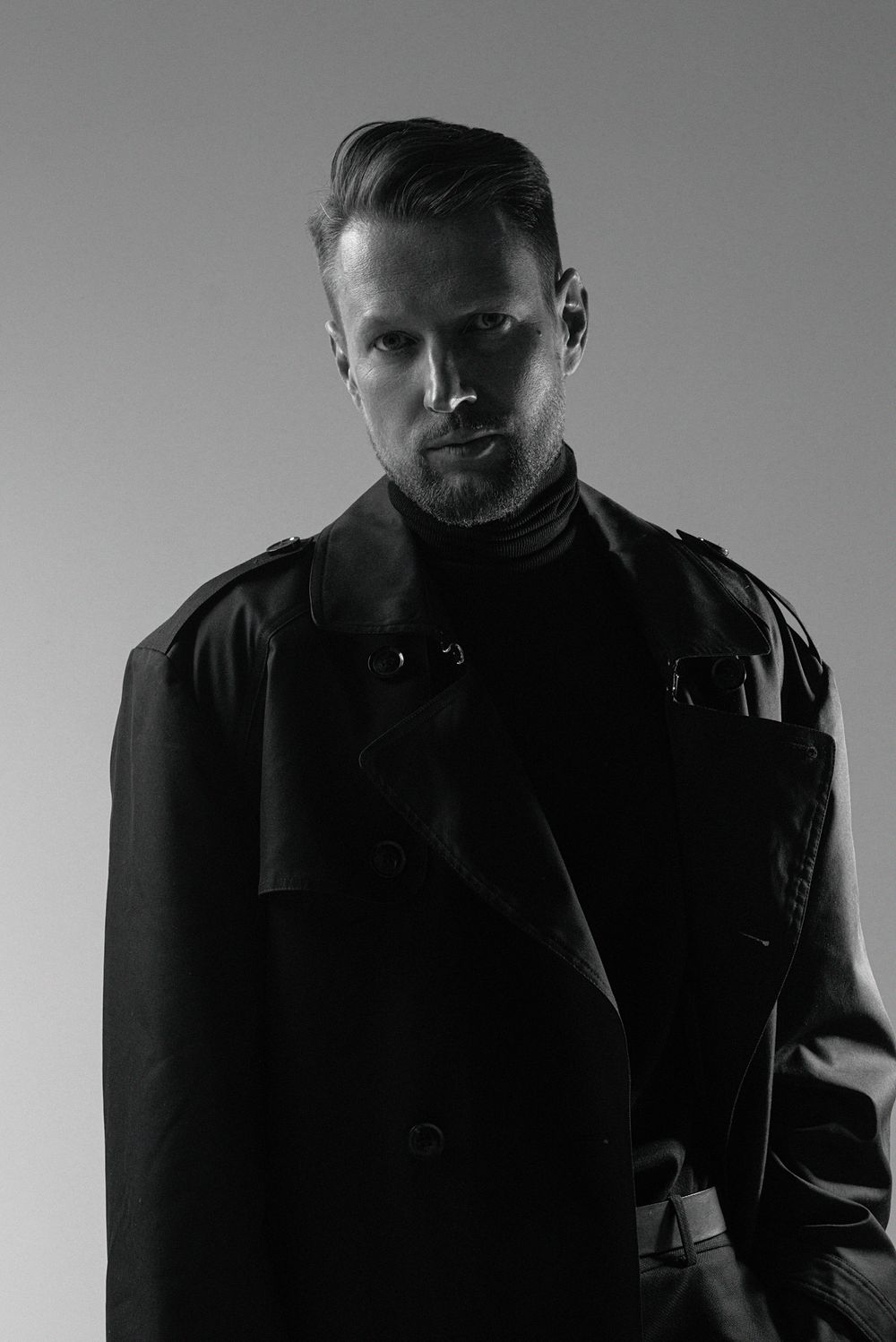 Dramatic black and white portrait showing figure in dark jacket with moody lighting and stern expression.