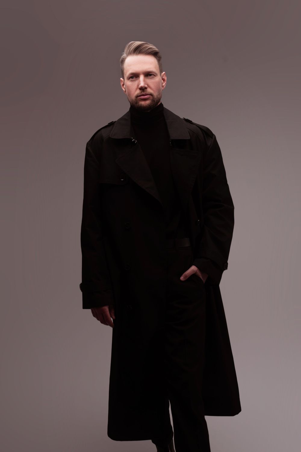 A person in an elegant black winter overcoat and turtleneck poses against a gray studio backdrop.