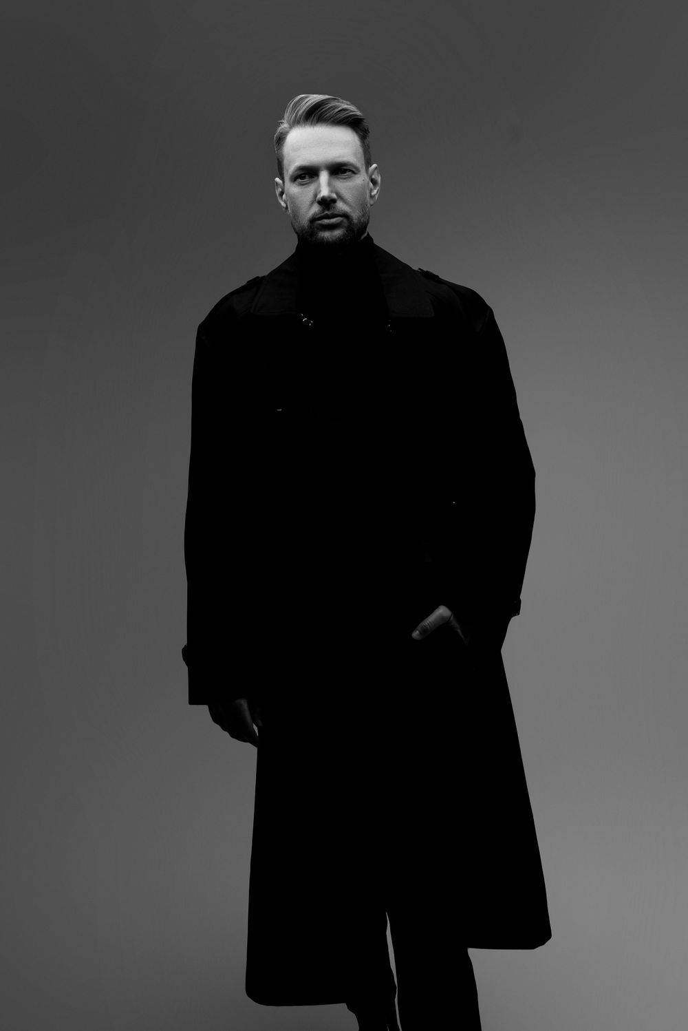 A dramatic black and white studio portrait of someone in a long dark winter coat against a grey gradient background.