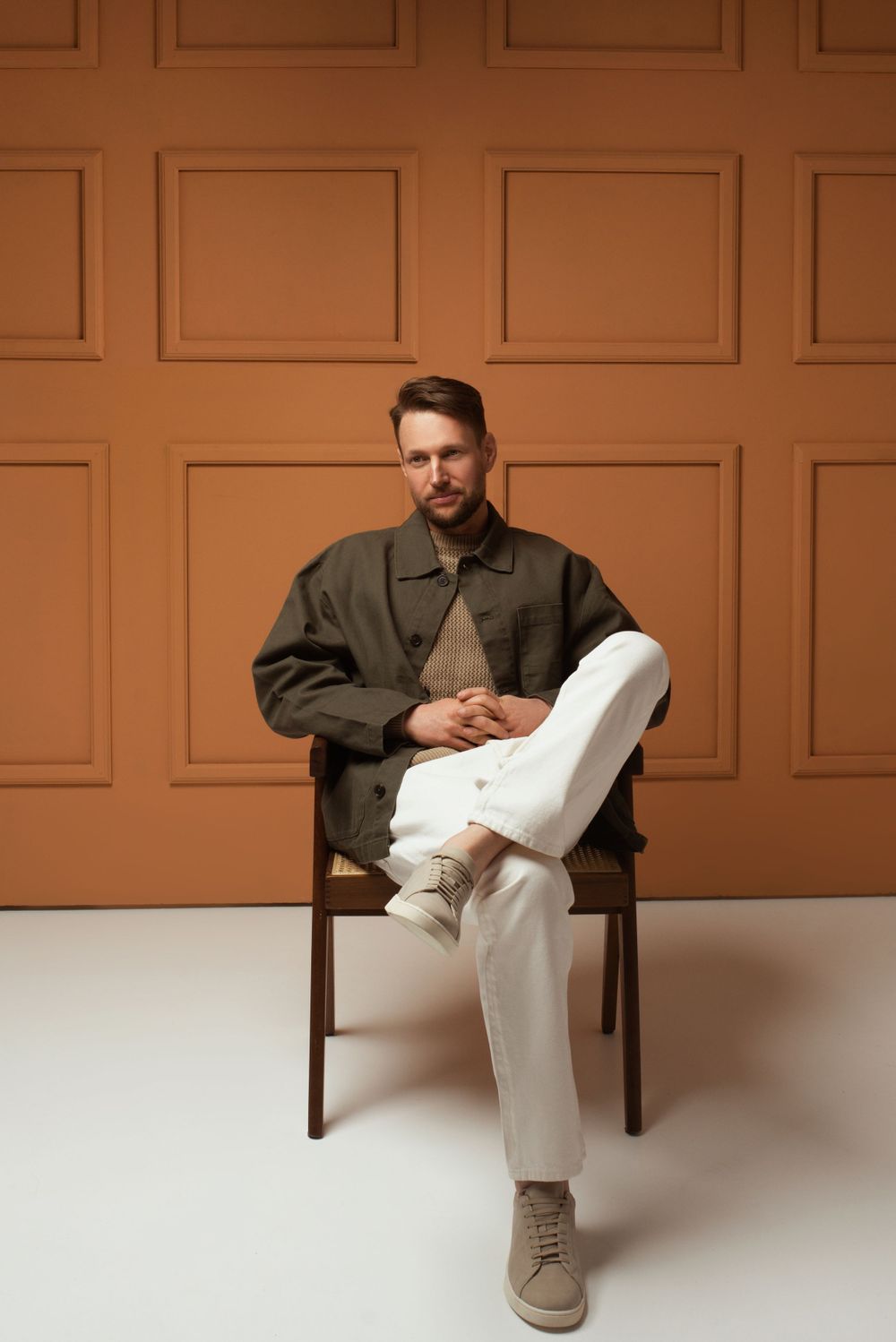 Person in olive jacket and white pants sits on wooden chair against orange paneled wall with modern minimalist styling.