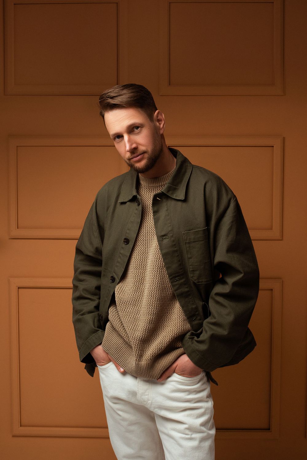 A person wearing an olive green jacket over a beige sweater and white pants poses against a brown textured wall.