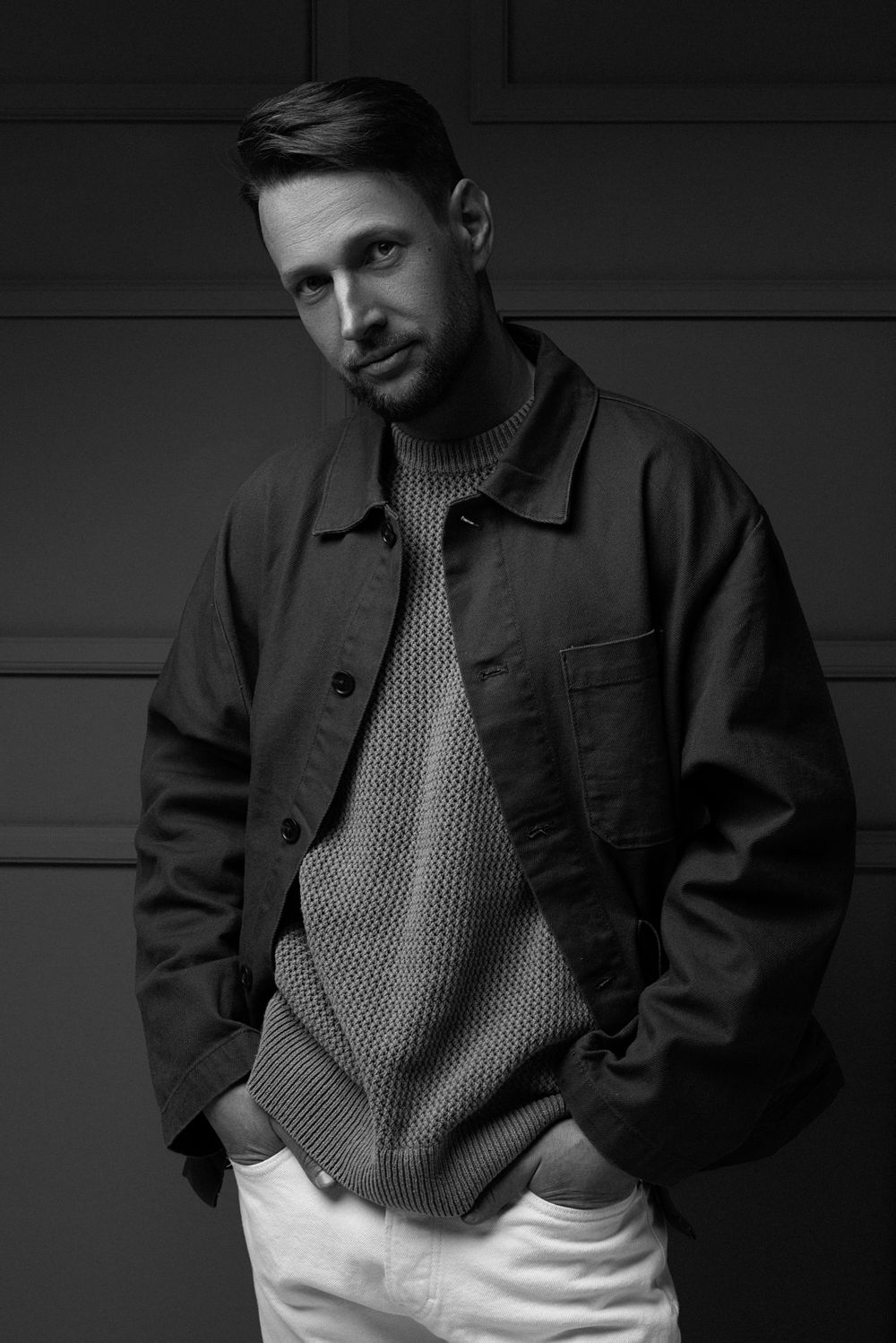 Black and white fashion portrait of someone in a casual dark jacket over knit sweater and white pants leaning against wall.