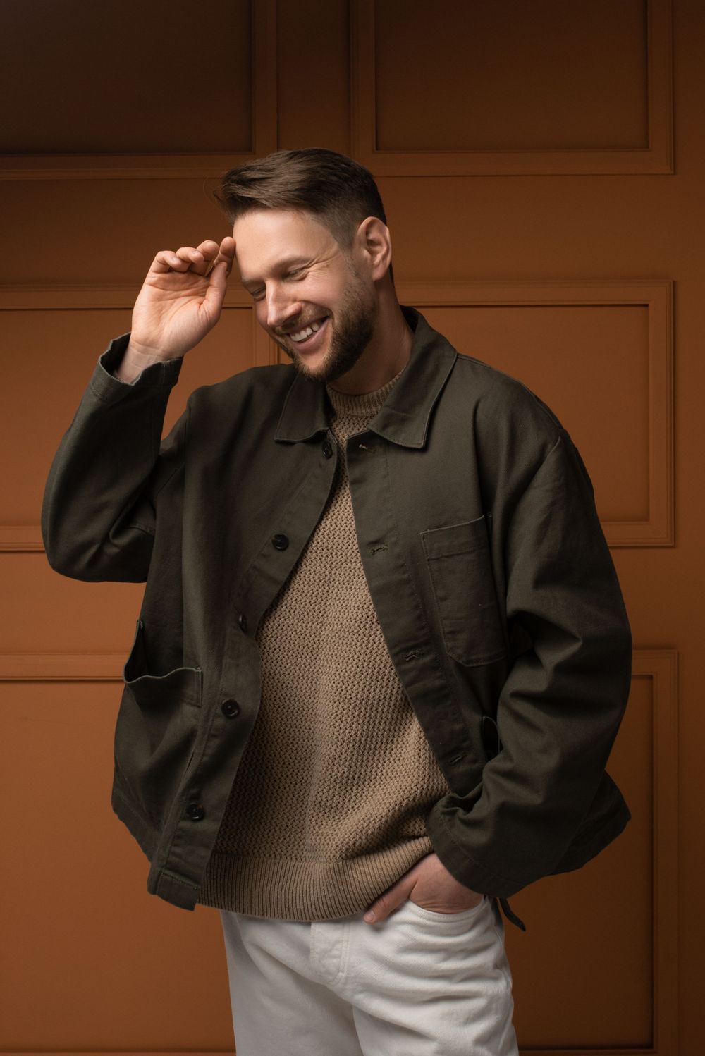 Stylish casual outfit featuring a dark jacket, beige sweater, and white pants against a brown background.