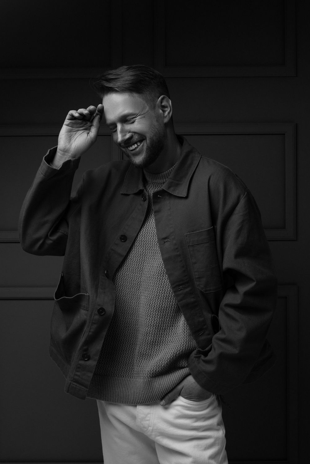 A black and white portrait of someone wearing a casual jacket and sweater, smiling while looking down.