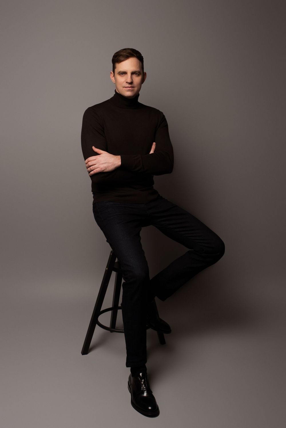 Professional studio portrait of someone in a black turtleneck sweater and dark pants seated on a wooden stool against grey backdrop.