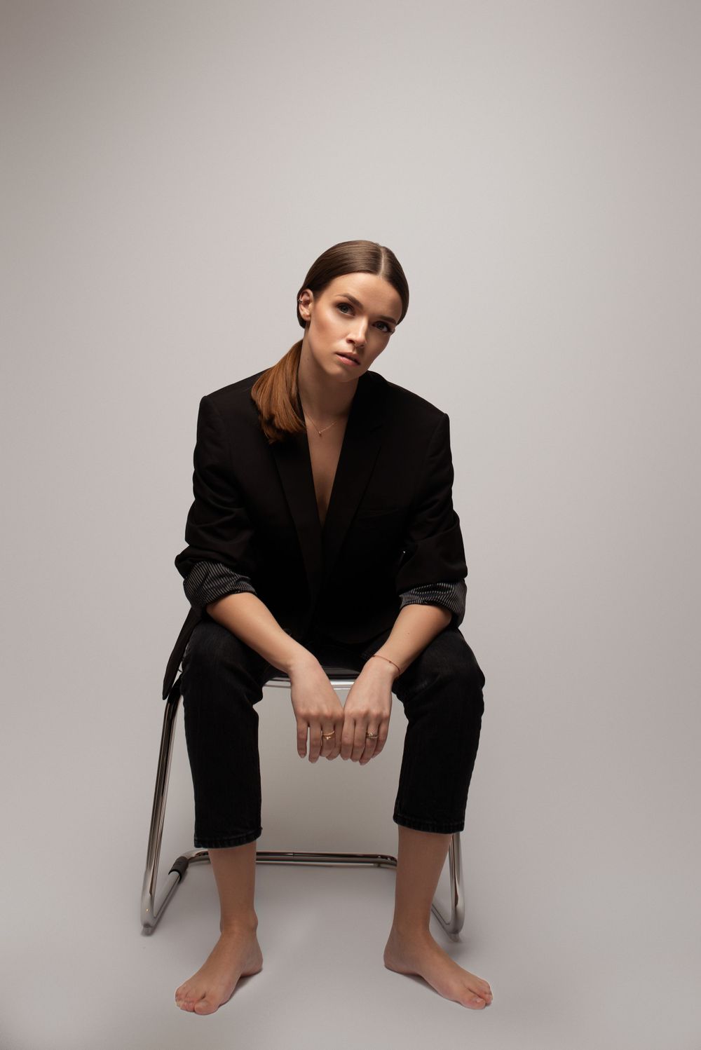 A model in a black oversized suit sits barefoot on a chair against a neutral backdrop in a minimalist fashion pose.