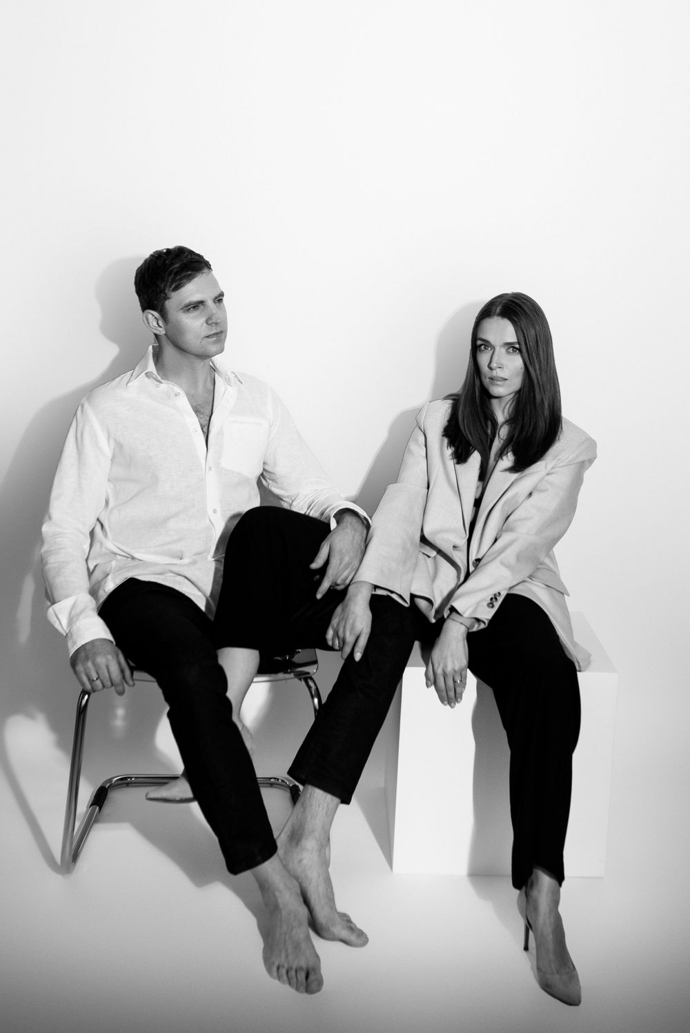 Two people in white shirts and black pants sit casually barefoot on chairs against a white background in black and white photo.