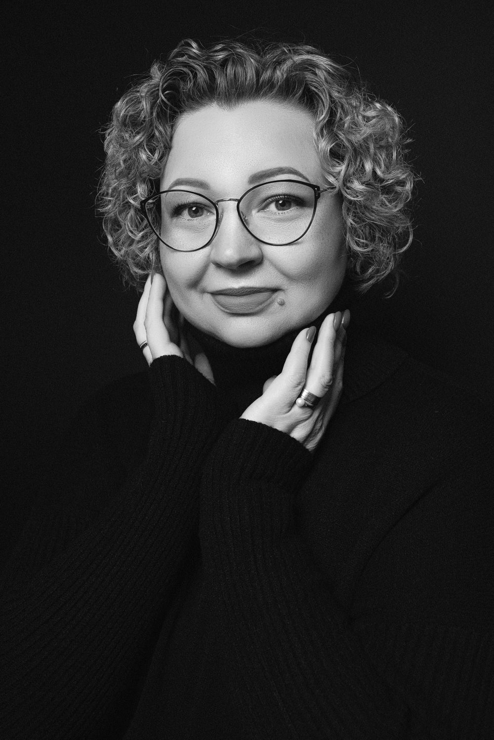 Black and white portrait of someone with curly hair and round glasses wearing a black turtleneck sweater.