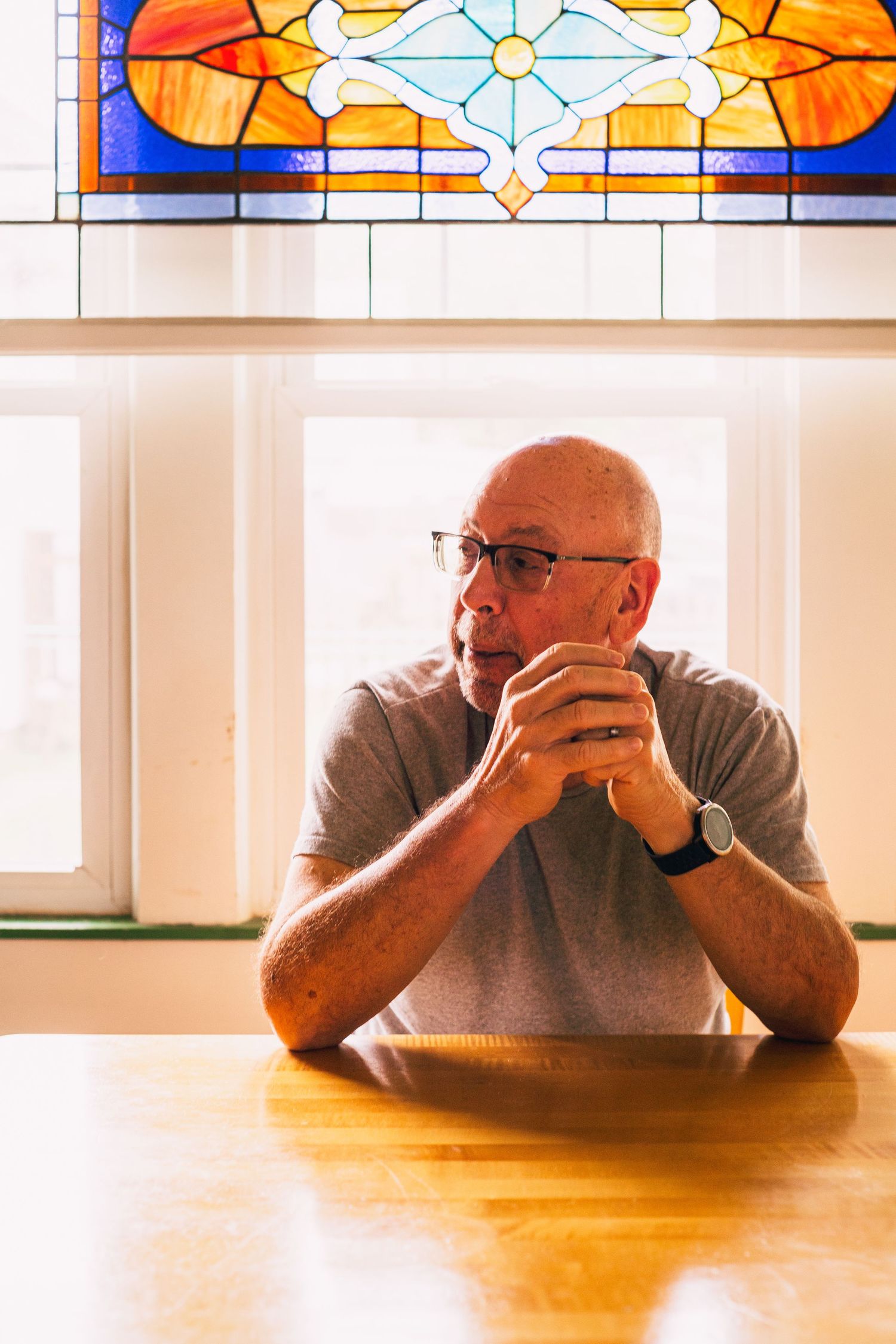 A person sits at a wooden table in contemplation beneath decorative stained glass windows.