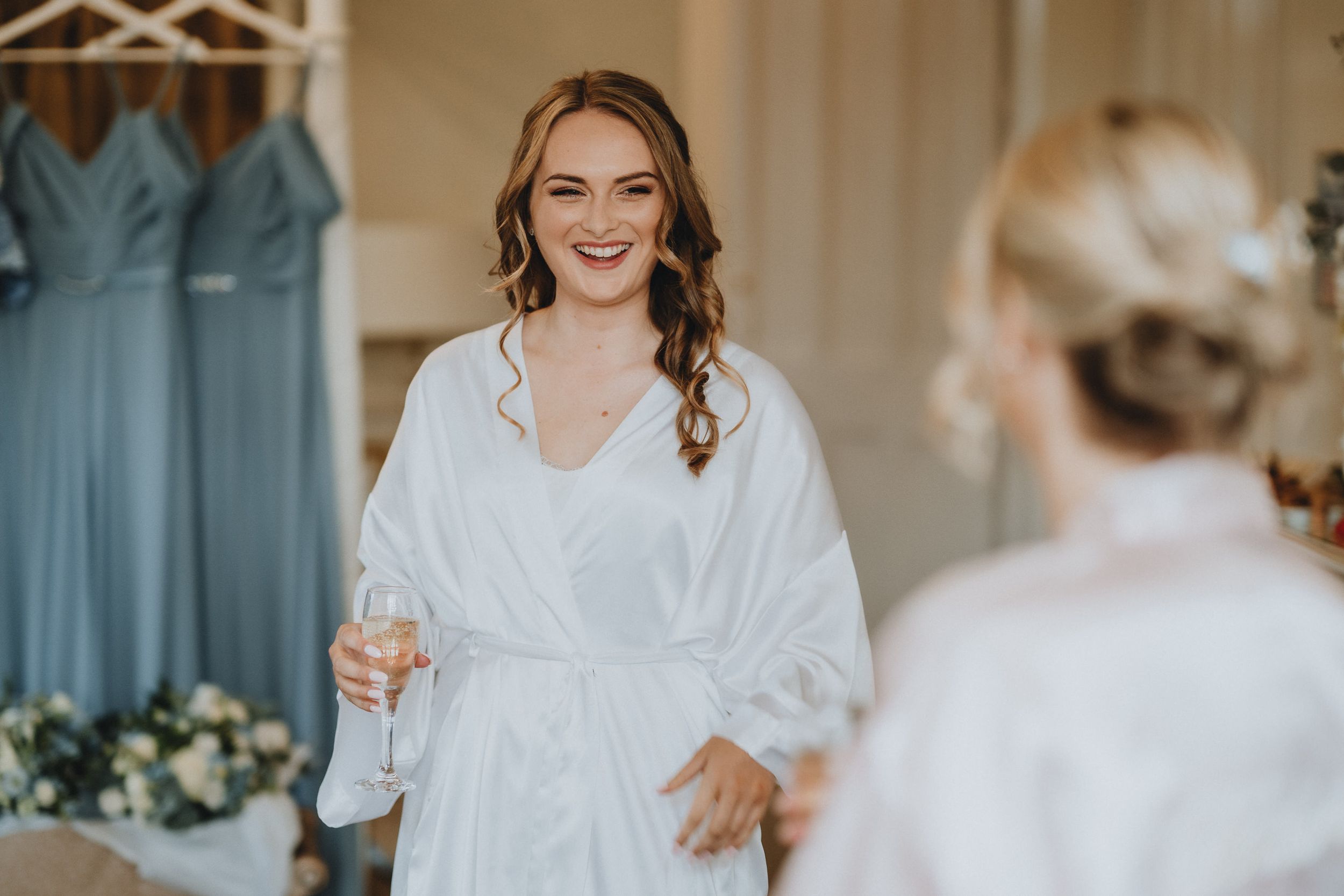 A person in a white robe smiles while holding champagne with blue bridesmaid dresses visible in the background.