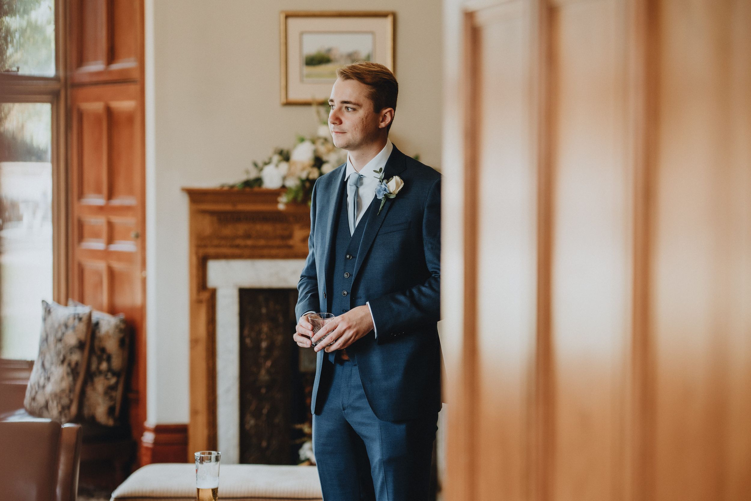 A person in a navy blue suit and tie stands pensively in an elegant room with a fireplace and soft lighting.