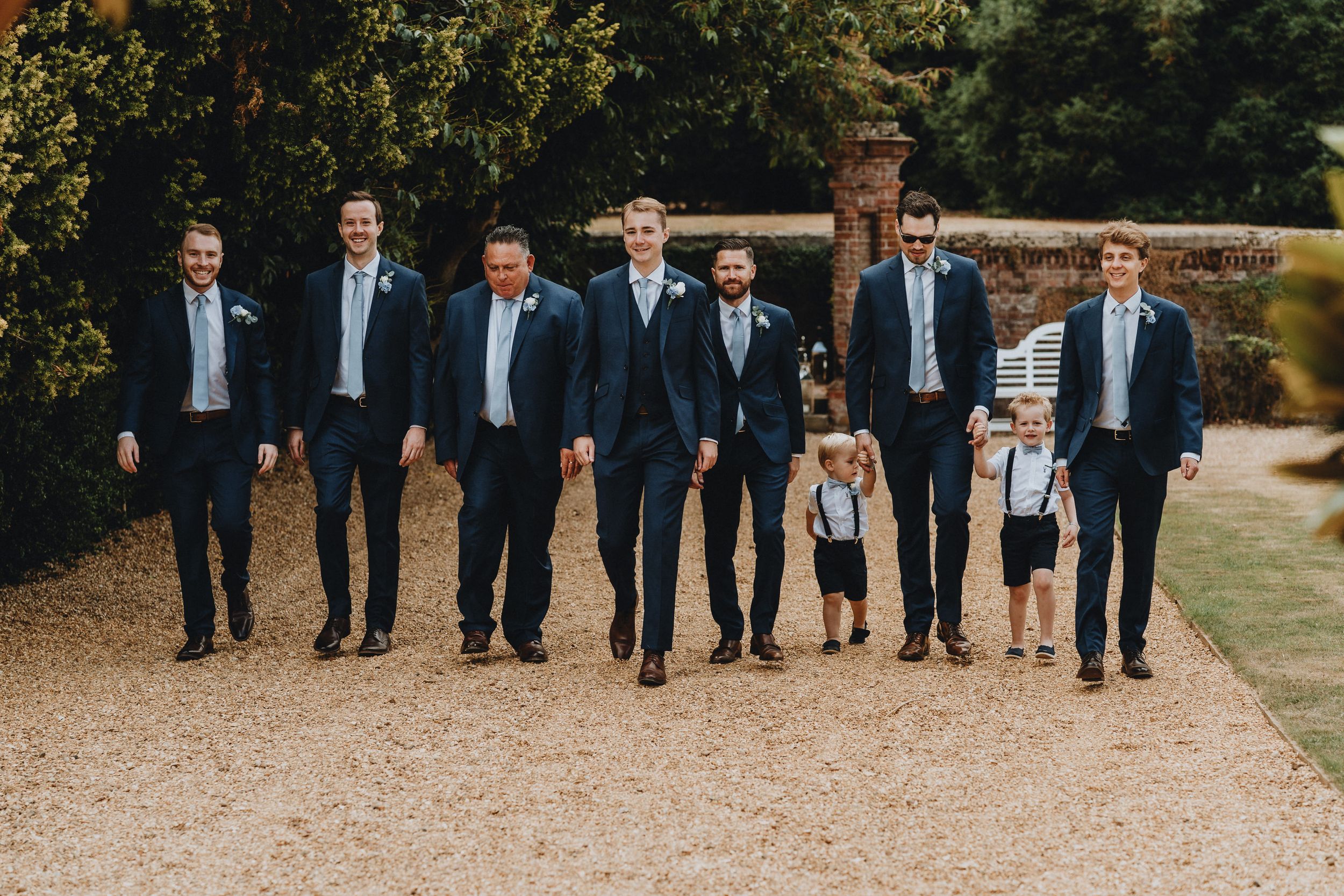 A wedding party group walks along a gravel path while wearing navy suits with a young ring bearer in suspenders and shorts.