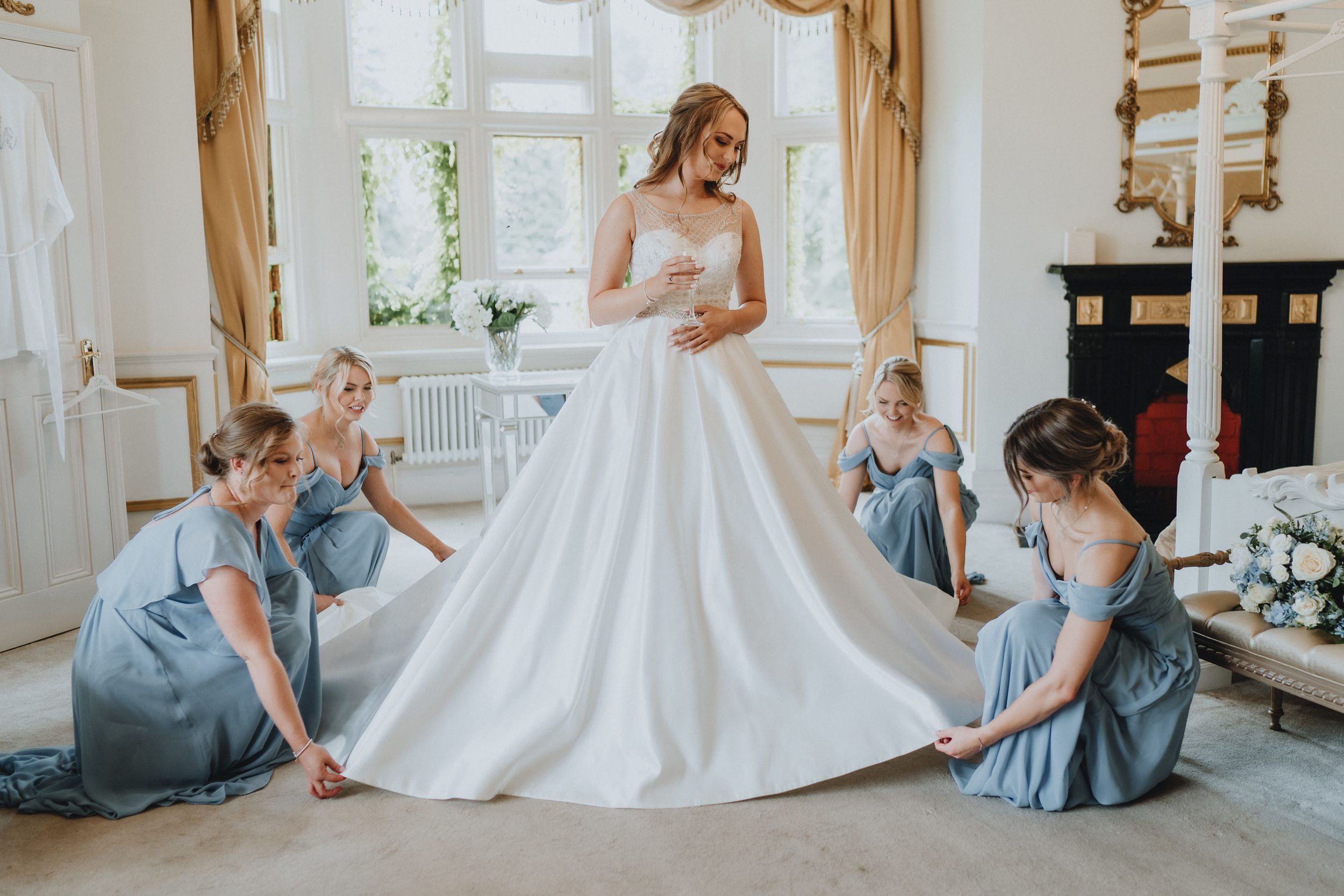 Bridesmaids in light blue dresses help adjust the flowing white wedding gown in an elegant room at Holmewood Hall, Peterborough.