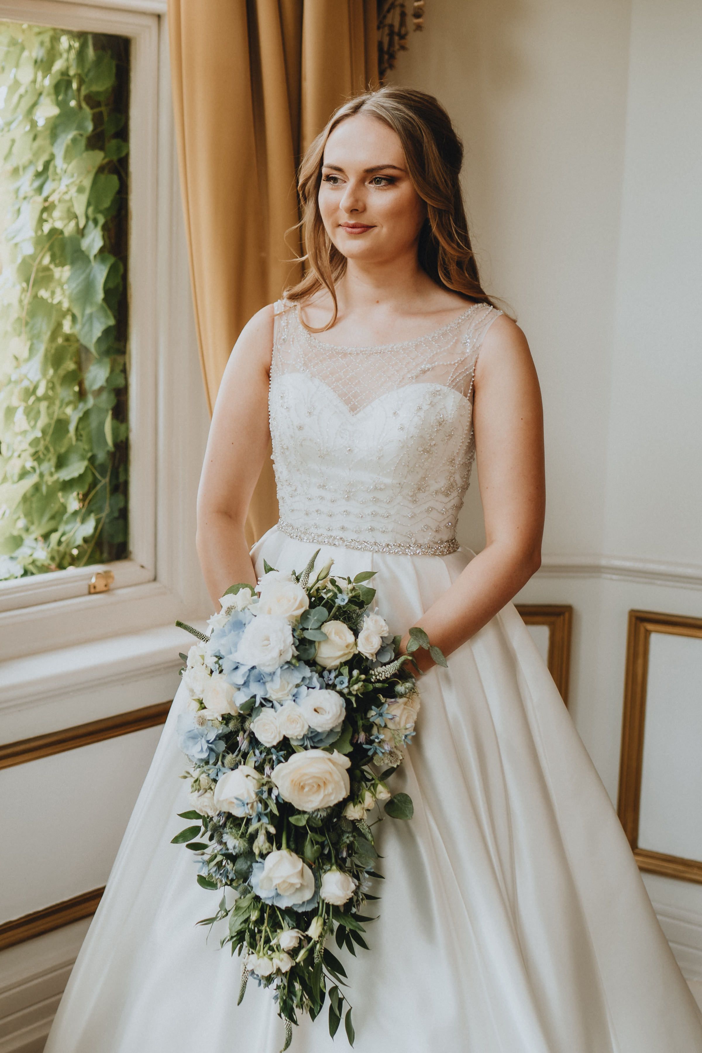 Bride in elegant white gown with illusion neckline holds cascading bouquet of white roses by sunny window with ivy.