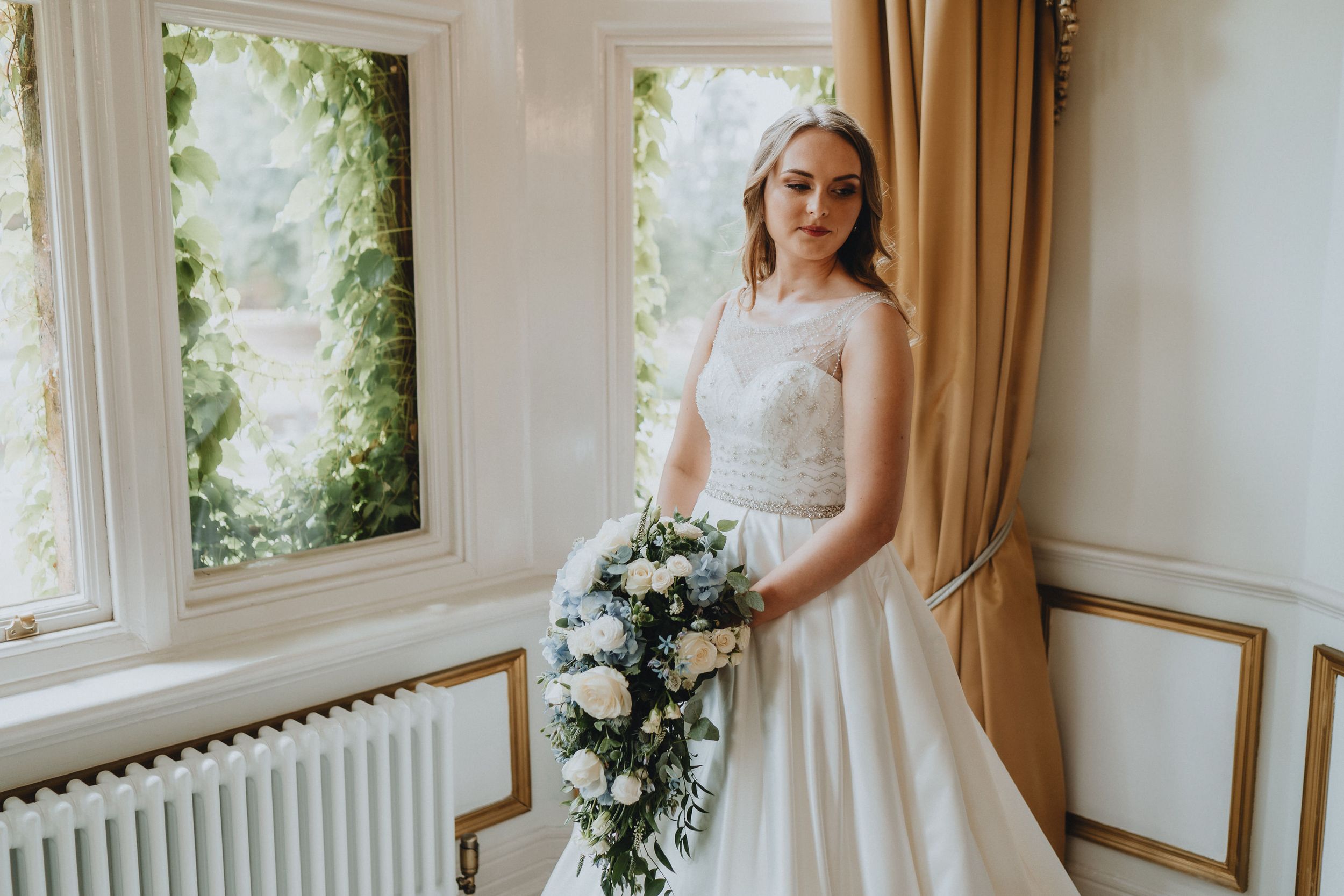 A bride in a white lace wedding gown holds a cascading white floral bouquet near sunlit windows and cream curtains.