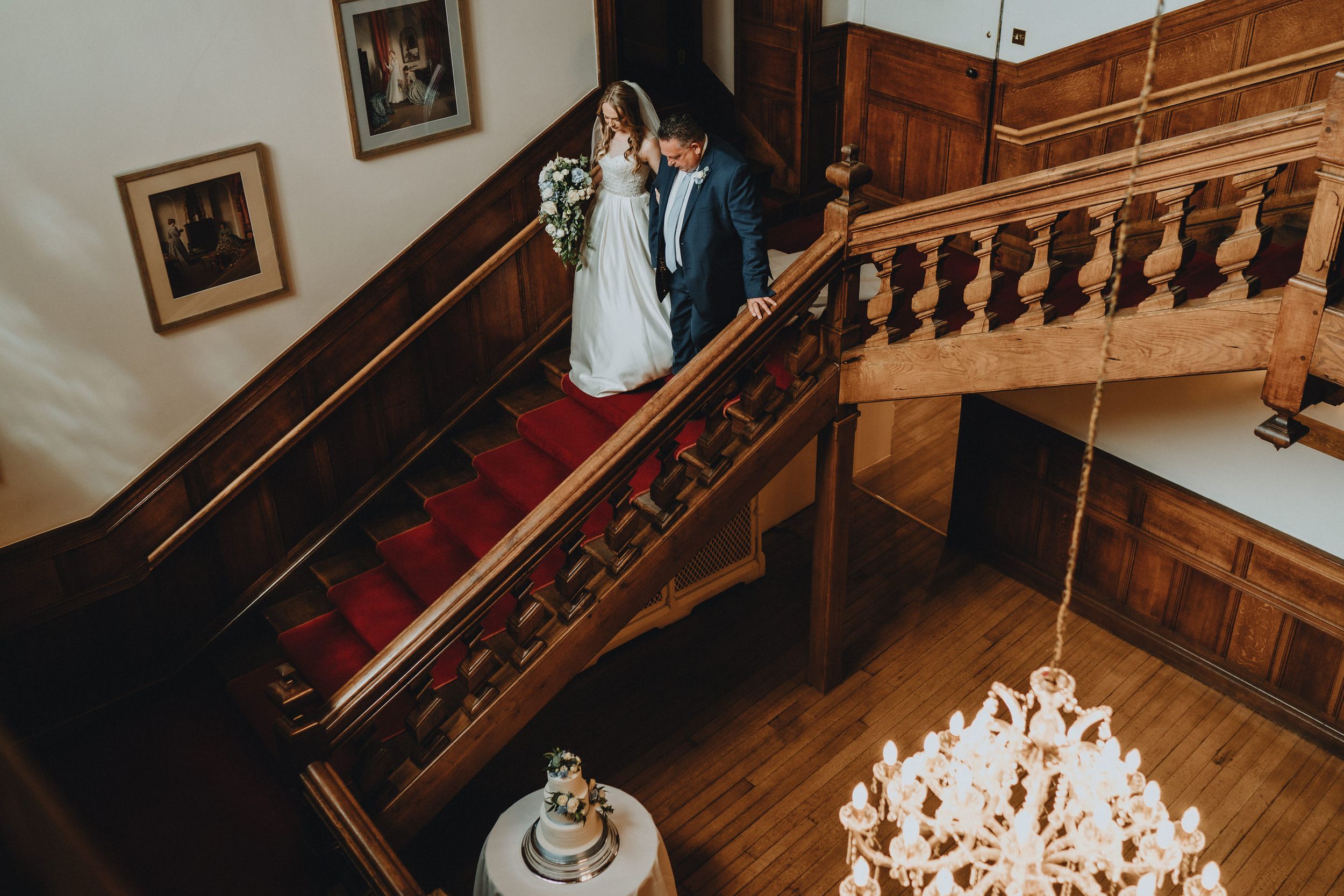 Bride and her father descends grand wooden staircase at Holmewood Hall with red carpet and chandelier in vintage mansion interior.