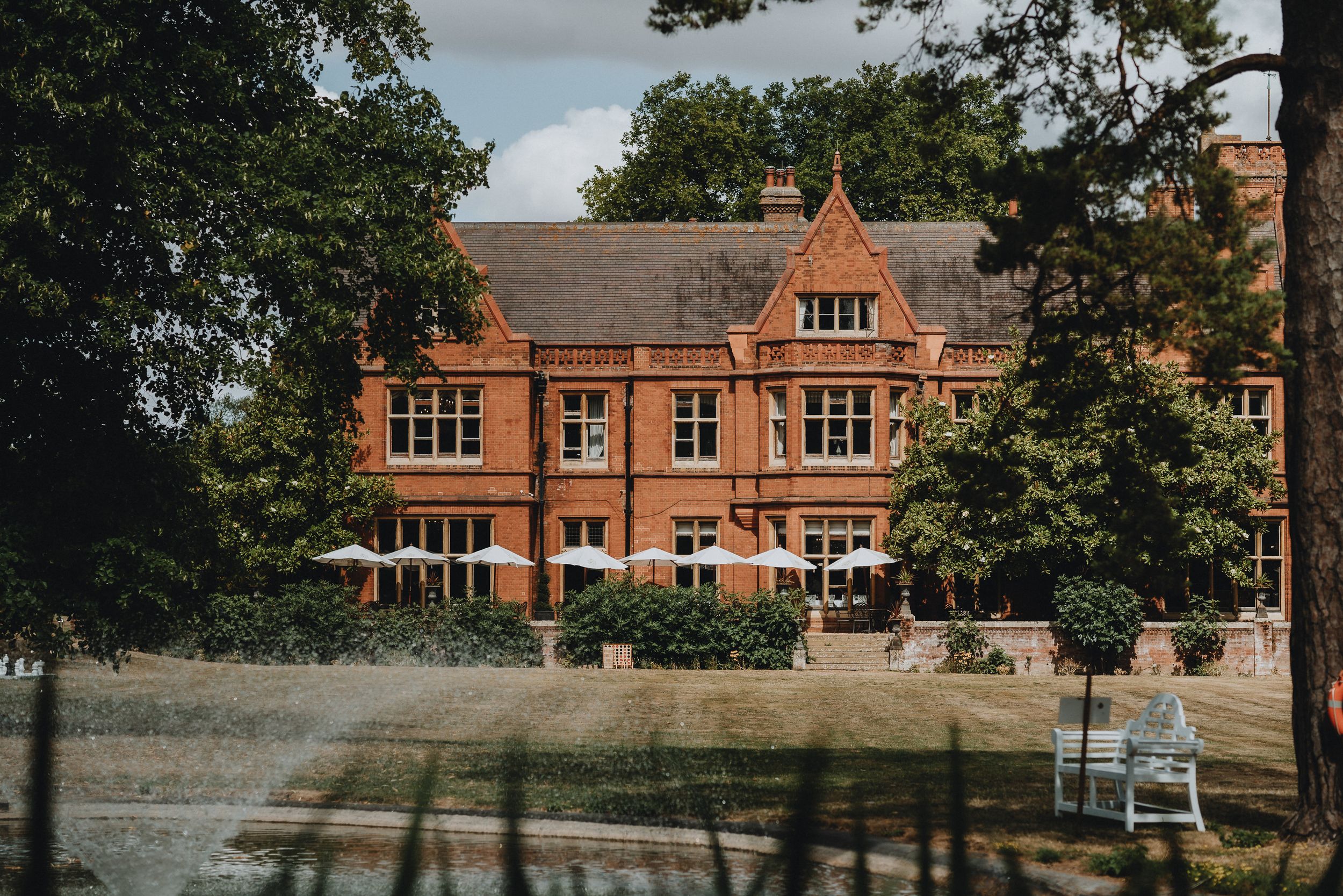Holmewood Hall a stately red brick Victorian mansion with gabled windows and dormer sits behind a manicured lawn on a sunny day.