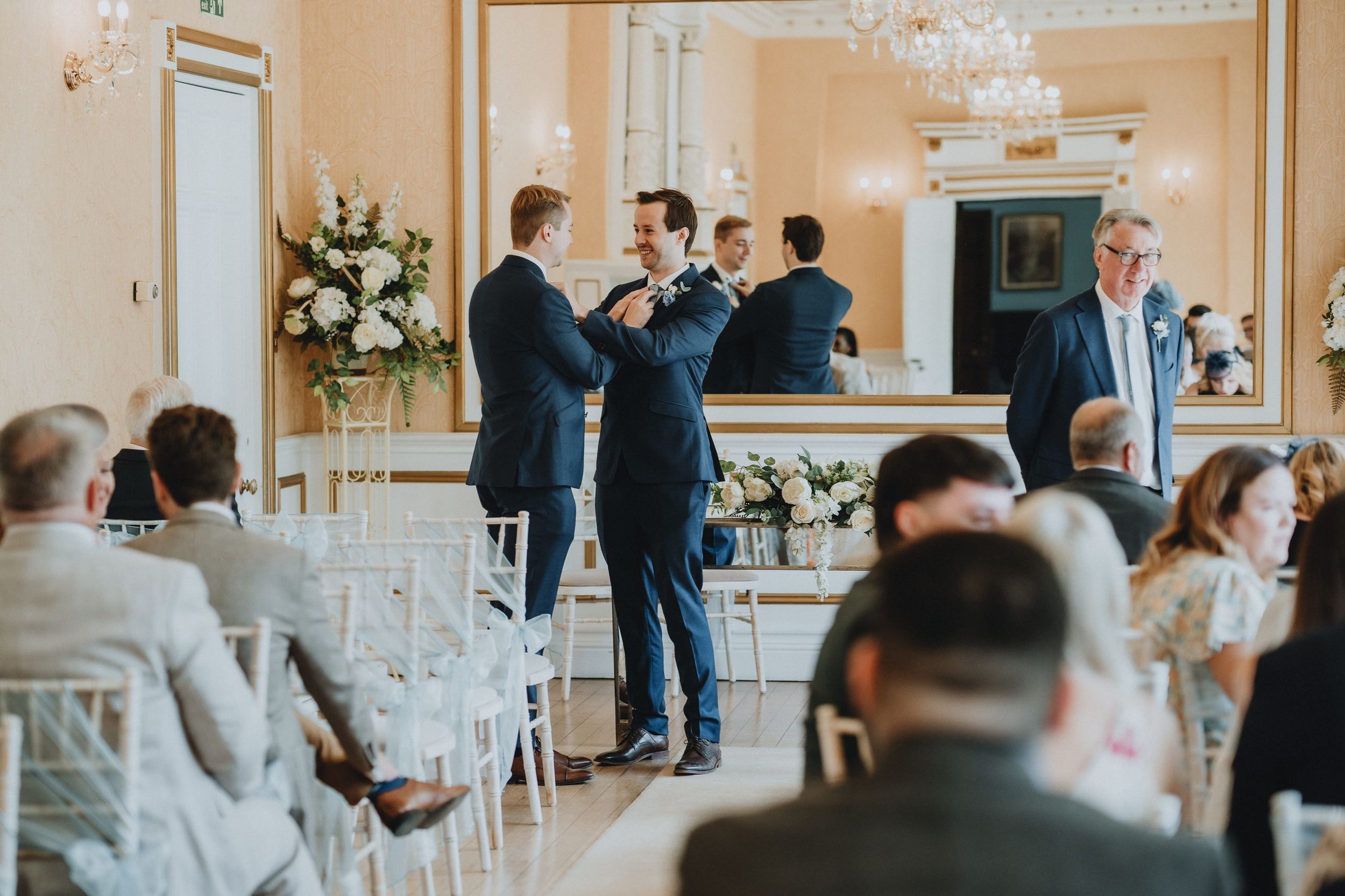 A wedding ceremony at Holmewood Hall takes place in an elegant room with white chairs and floral arrangements by a fireplace.