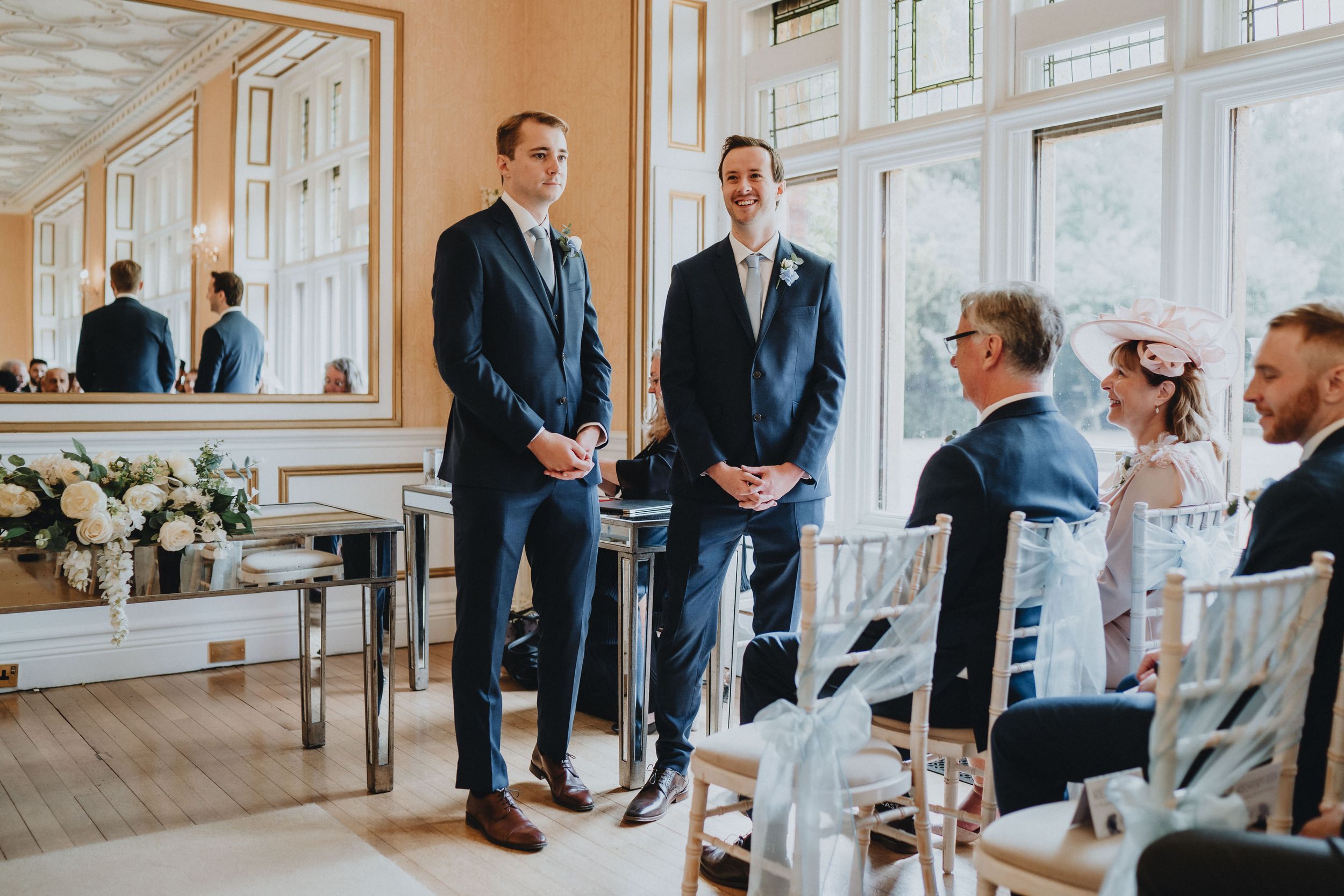 A wedding venue interior with hardwood floors, mirrors, and floral arrangements as attendees gather before the ceremony.