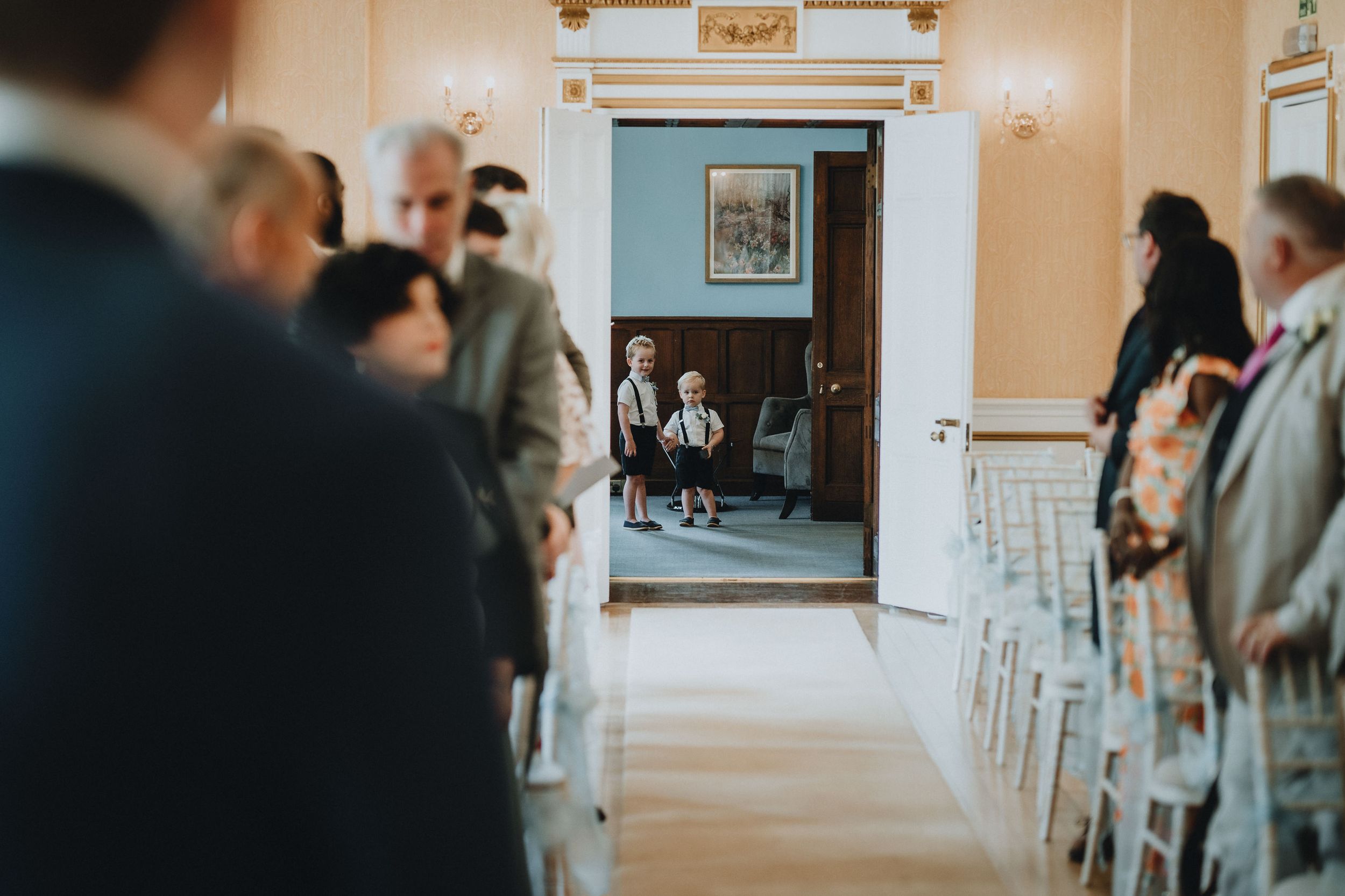 Two young figures in traditional attire sit in a doorway while wedding guests gather in a bright indoor venue.