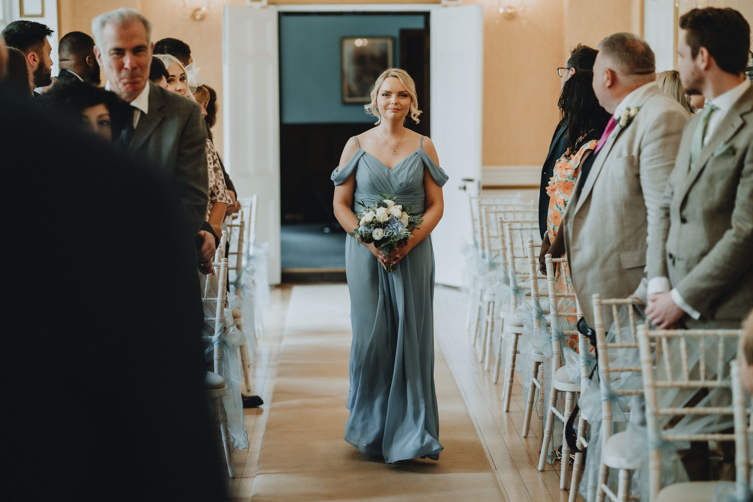 A wedding party member in a flowing blue gown walks down the aisle during a ceremony surrounded by guests.
