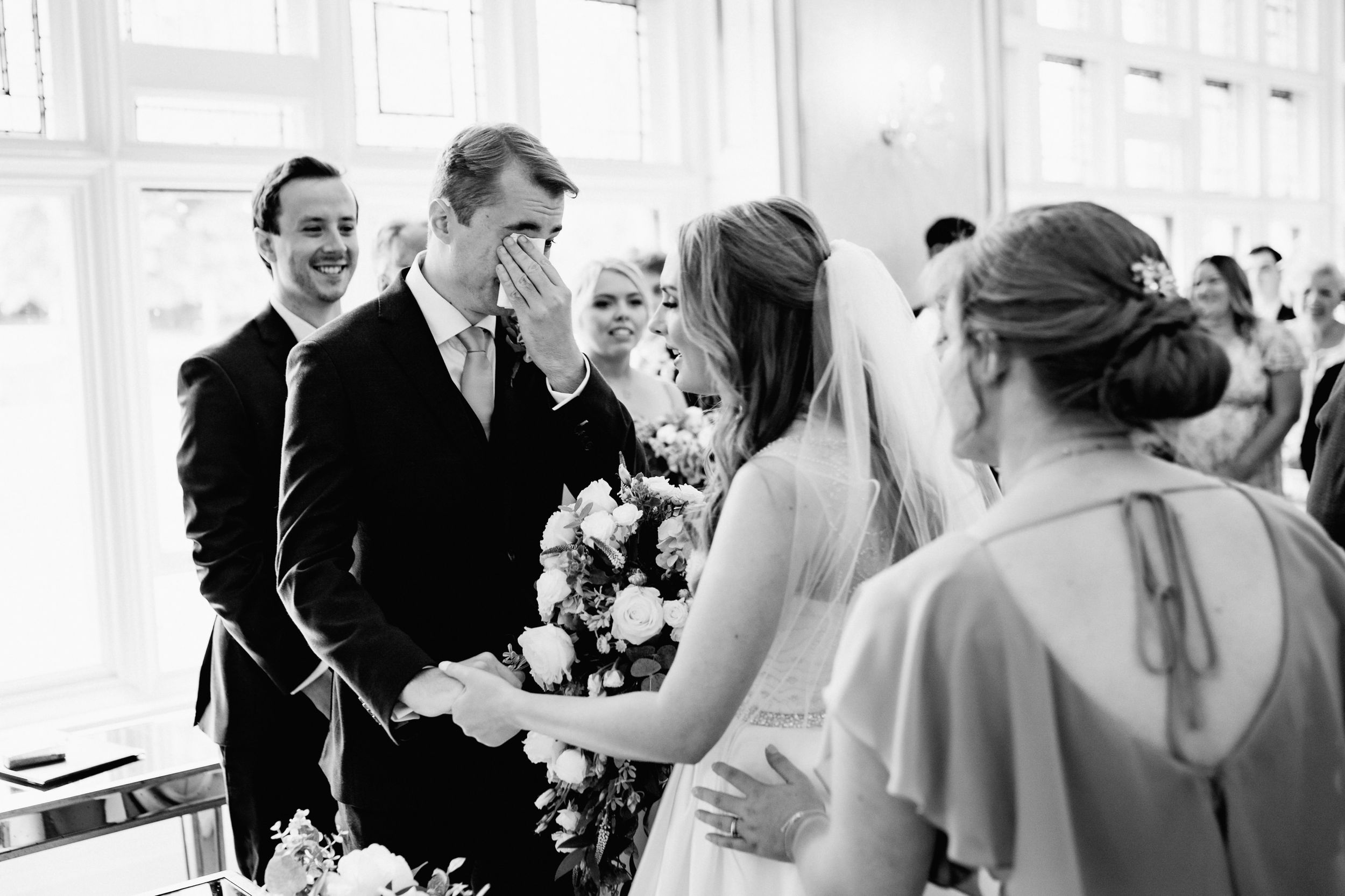 A black and white photo capturing an intimate moment during a wedding ceremony in a bright indoor venue.