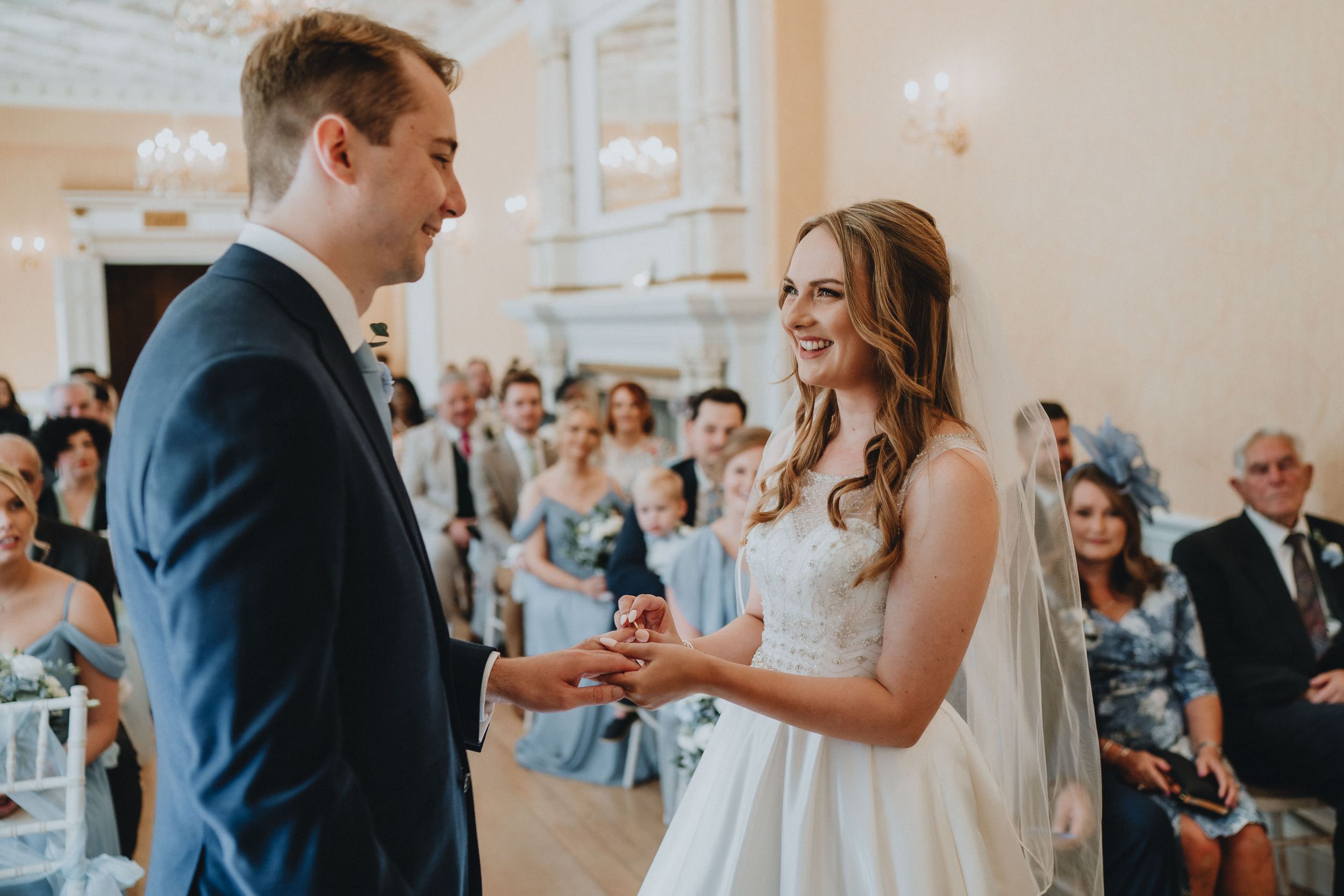 A couple exchanges rings during their wedding ceremony while guests look on in a bright, elegant venue.
