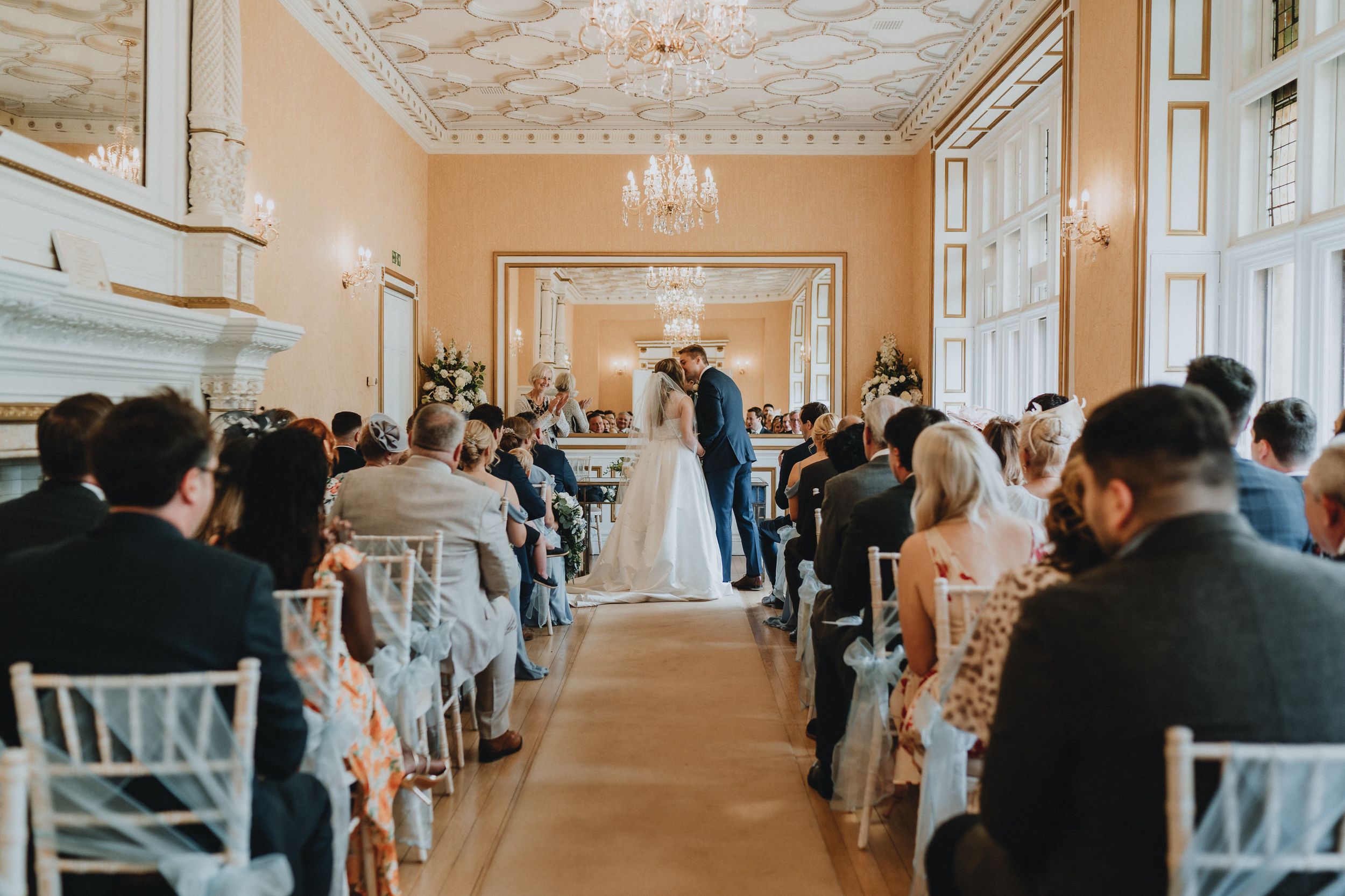 A wedding ceremony at Holmewood Hall takes place in an elegant room with ornate ceiling details and white chairs lining the aisle.