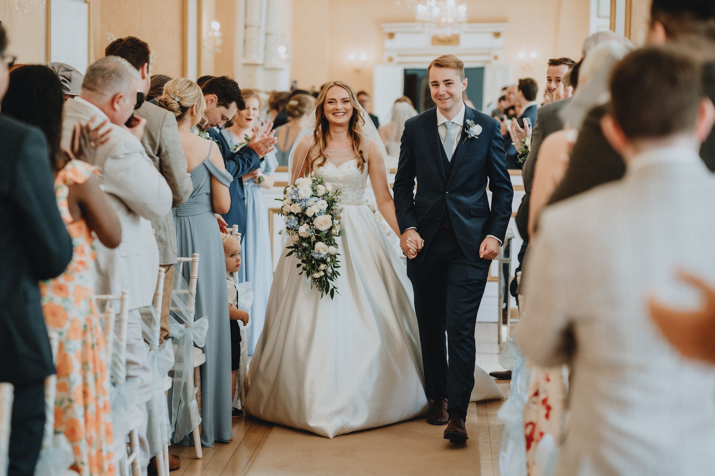 A bride and groom walk down the church aisle lined with guests, as natural light fills the ceremony space.