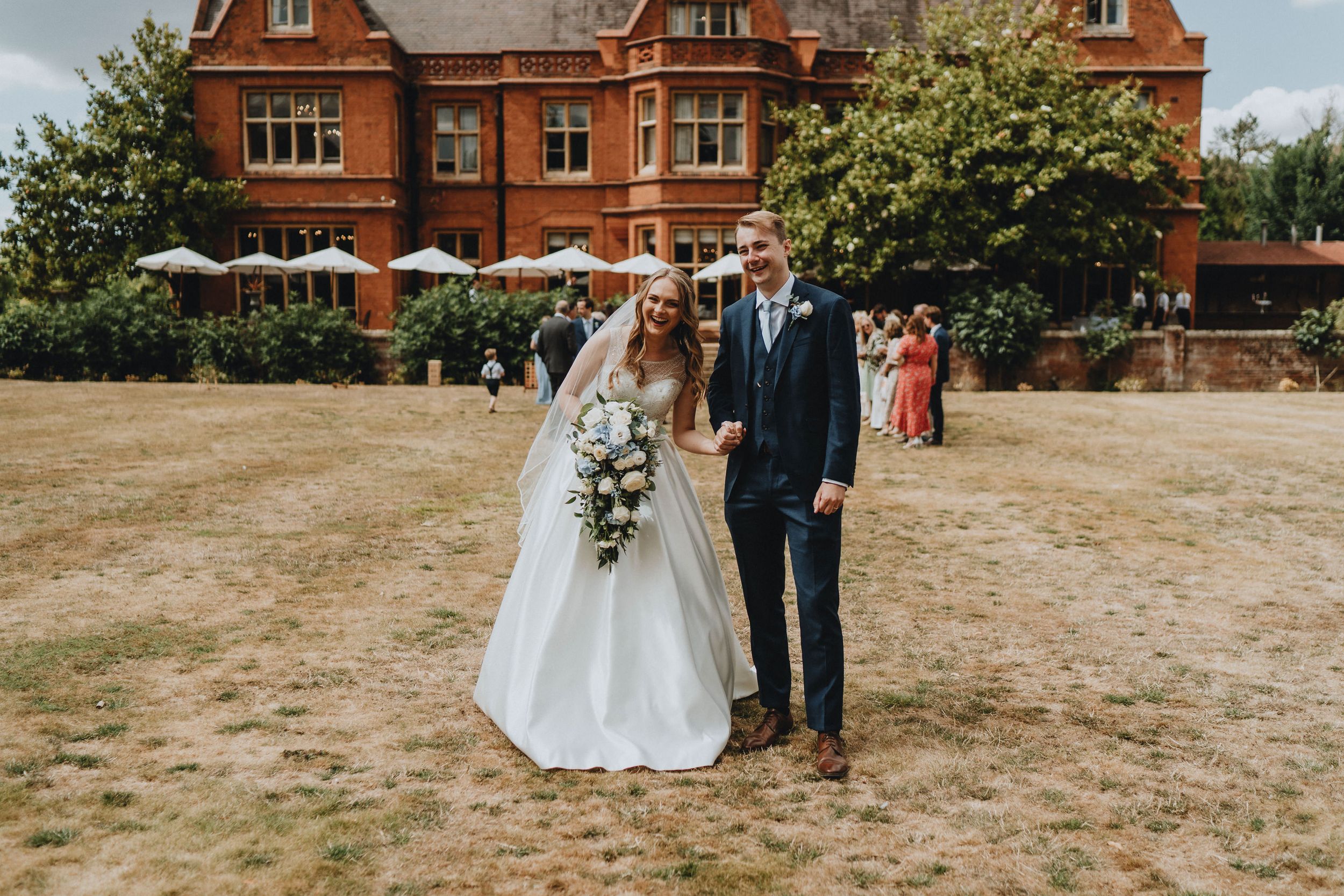 A wedding couple stands together on dried grass in front of a grand red brick Victorian mansion on a summer day.