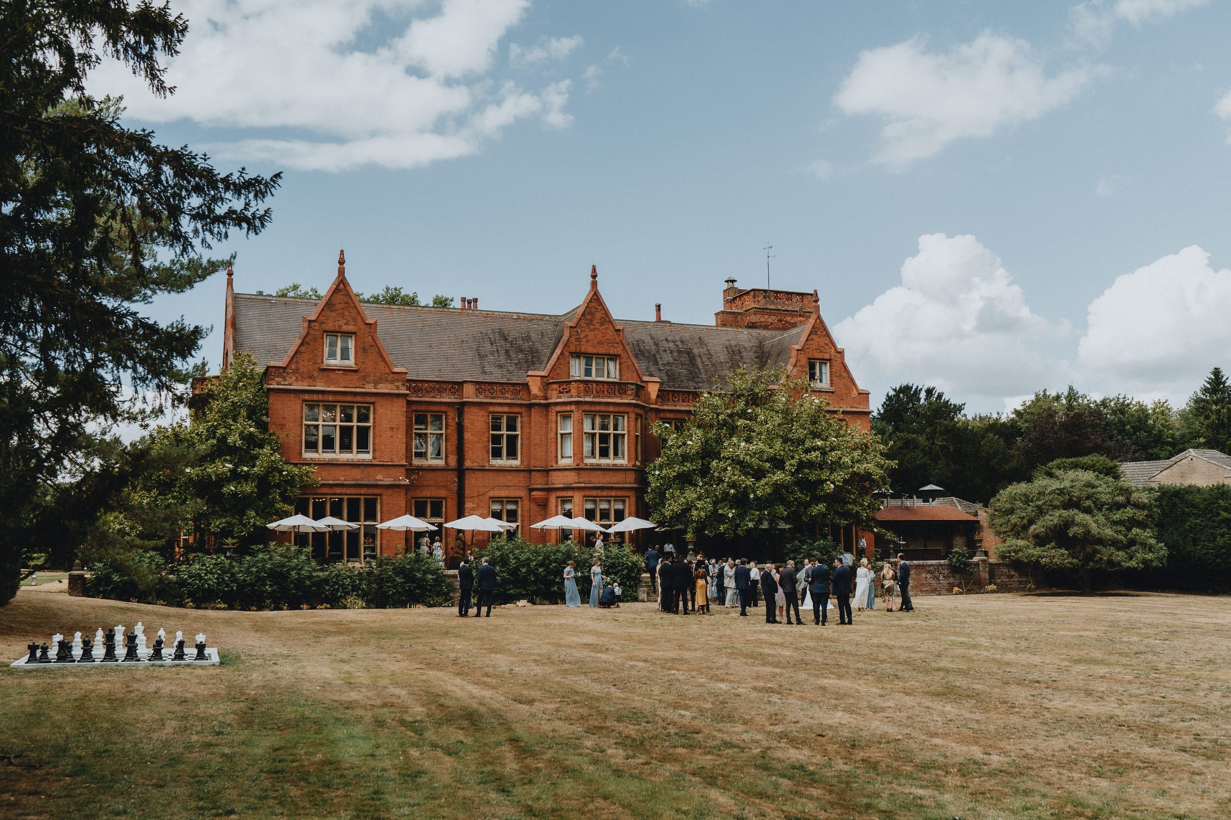 A large Victorian red brick mansion of Holmewood Hall with guests gathered outside on the lawn on a sunny summer day.
