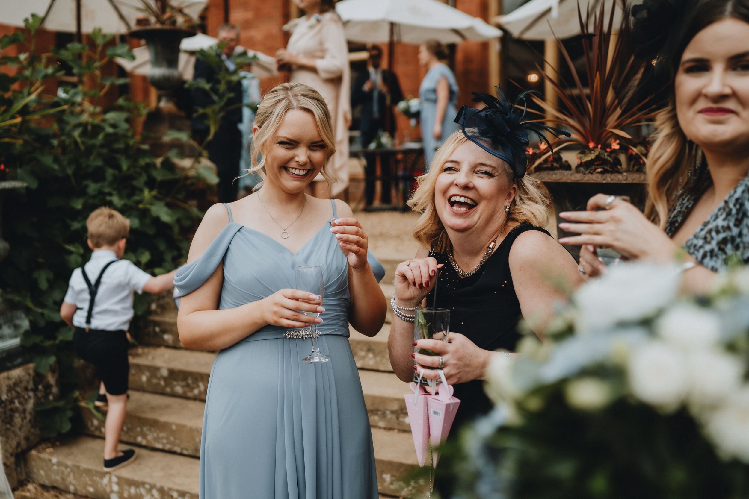 Wedding guests laughing and holding drinks at an outdoor garden reception with flowers and brick backdrop.