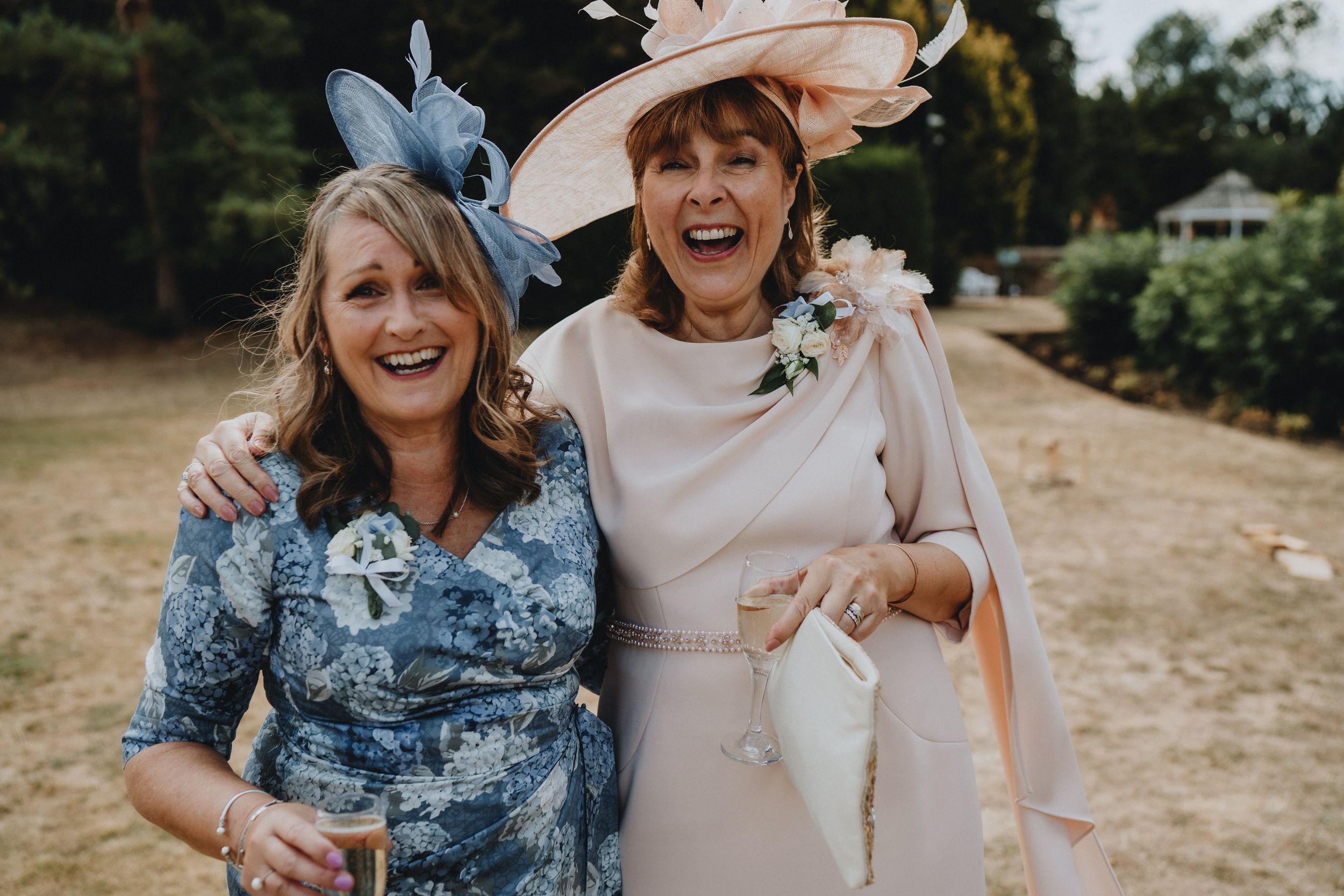 Wedding guests in elegant floral dresses and fancy hats sharing a joyful moment at an outdoor celebration.