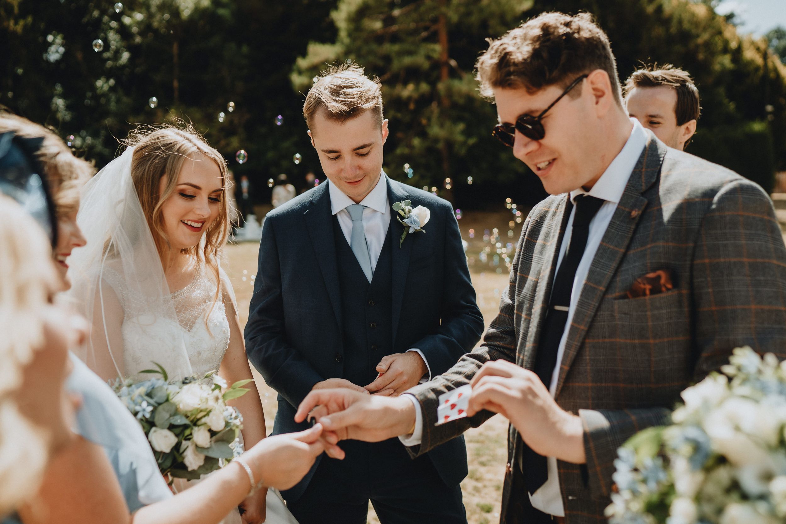 Wedding ceremony attendees exchange rings outdoors on a sunny day surrounded by floral arrangements.