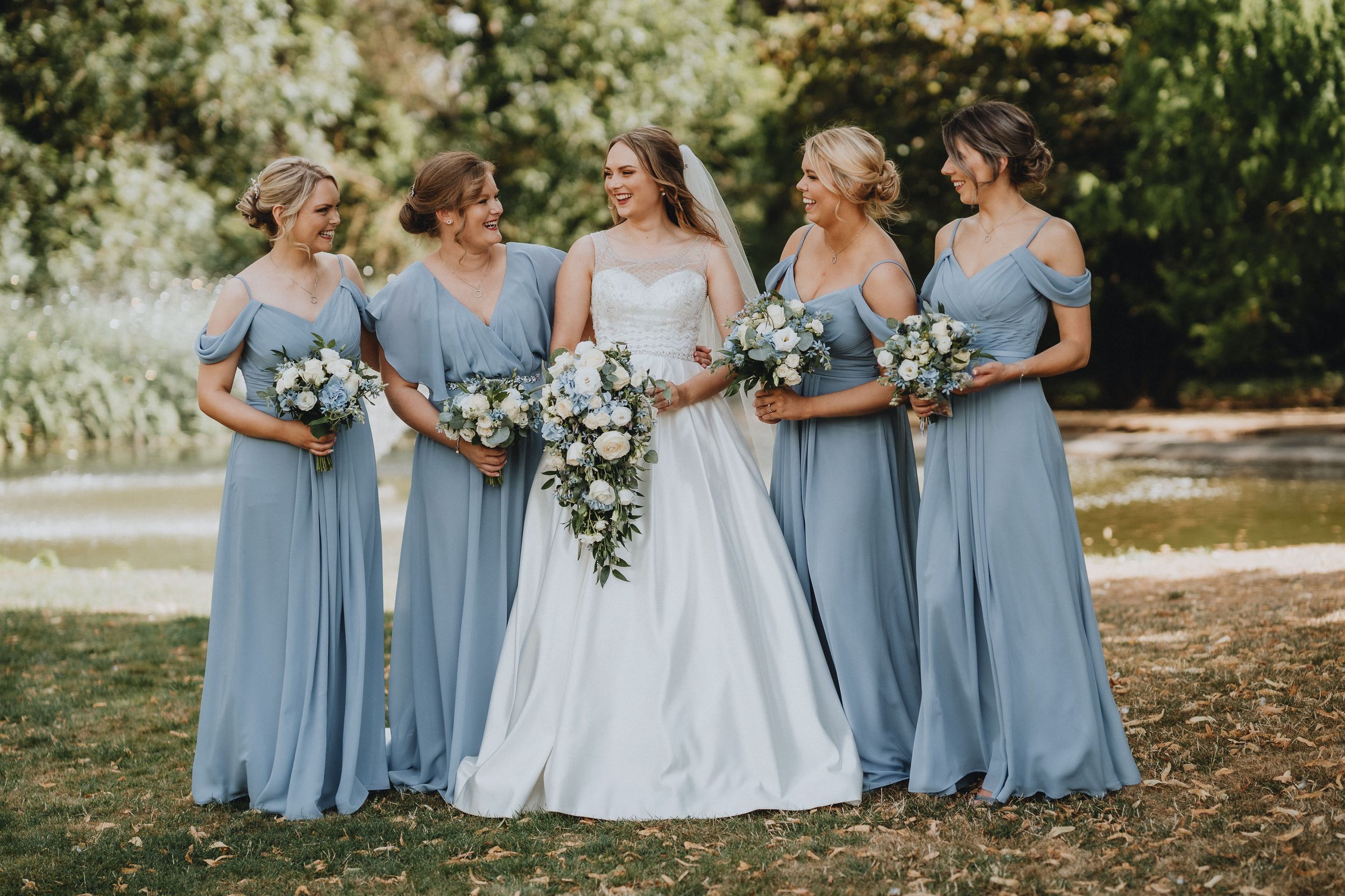 A wedding party in dusty blue bridesmaid dresses and white bridal gown pose together holding floral bouquets.
