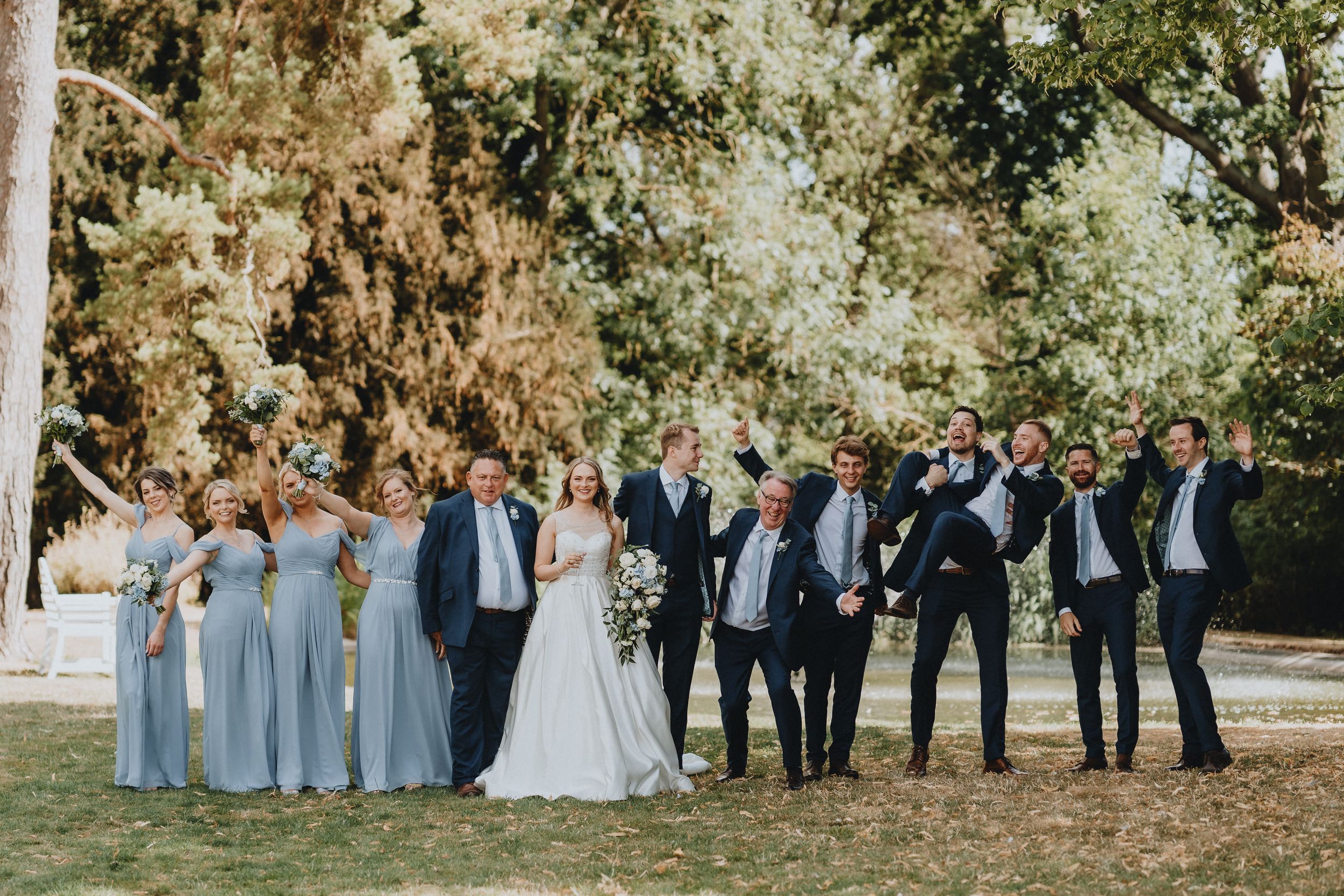Wedding party poses outdoors at Holmewood Hall in elegant attire with bridesmaids in light blue dresses and groomsmen in dark suits.