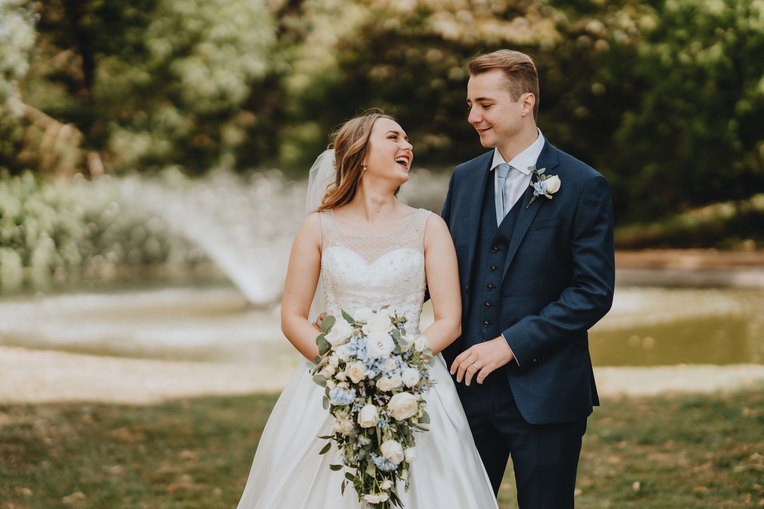 Couple in wedding attire laughing together outdoors while holding elegant white and green floral bouquet.