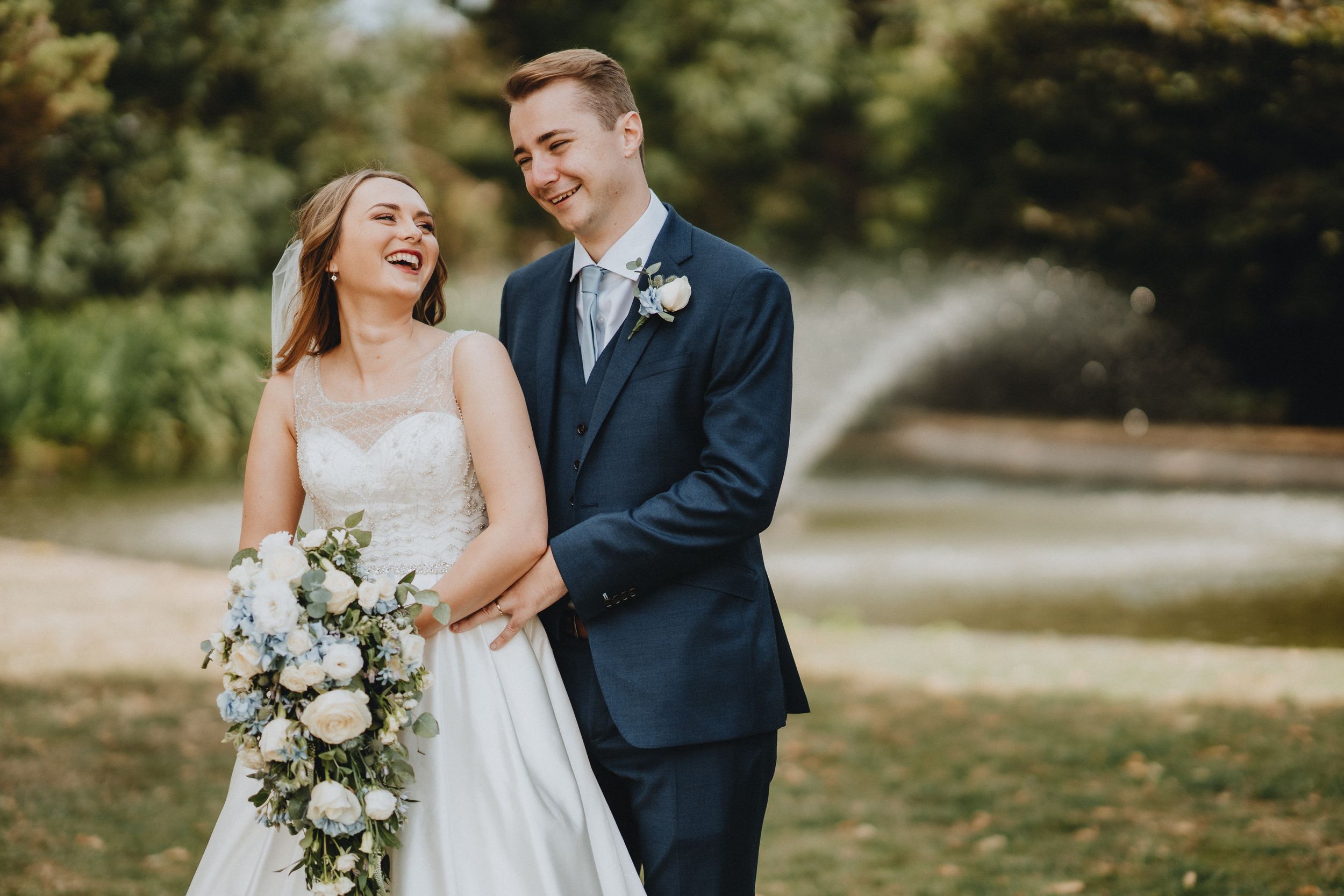 A joyful wedding couple laughing together outdoors while holding a white and green floral bouquet.