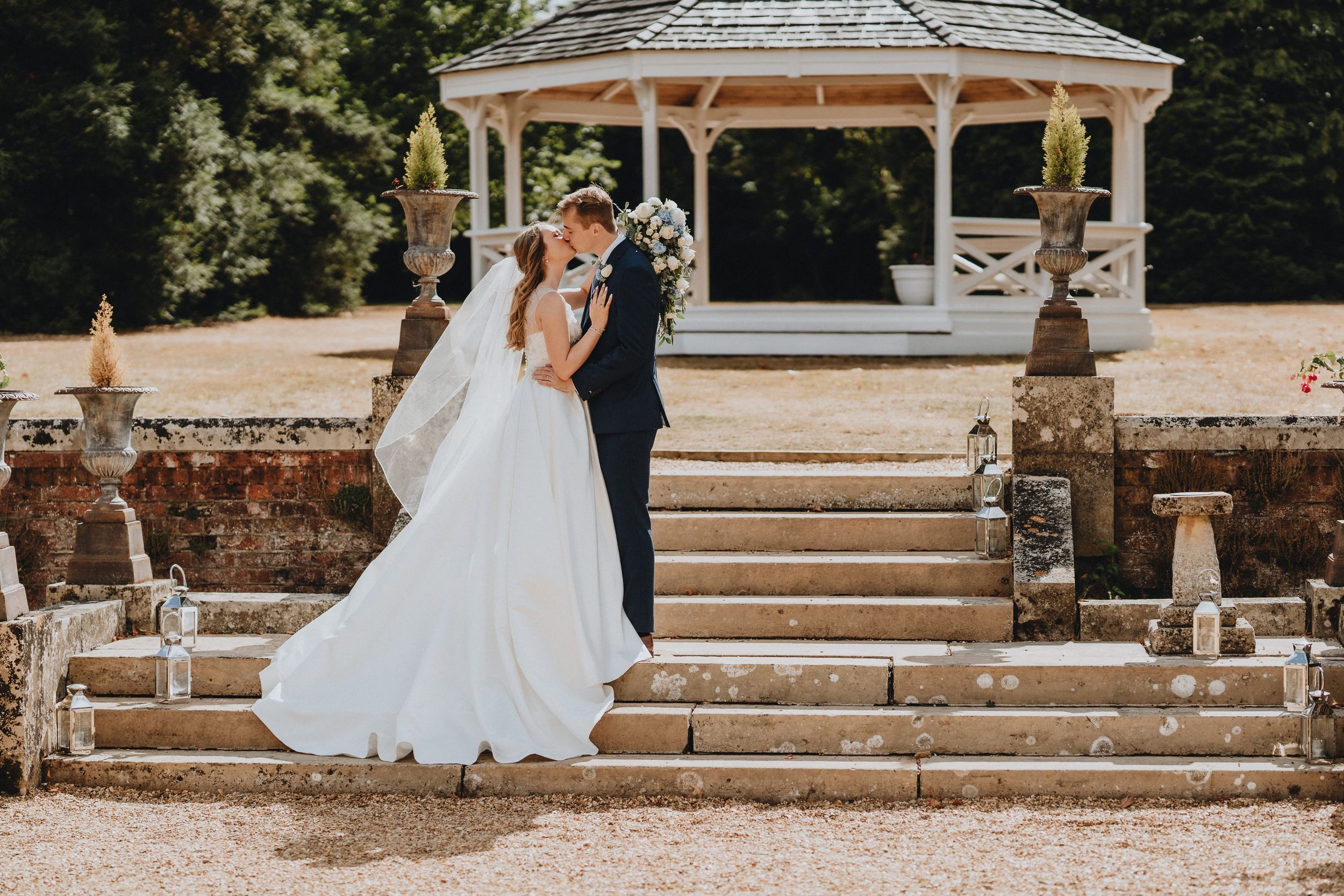 A wedding couple stands on stone steps in front of a white wooden gazebo in a garden setting.