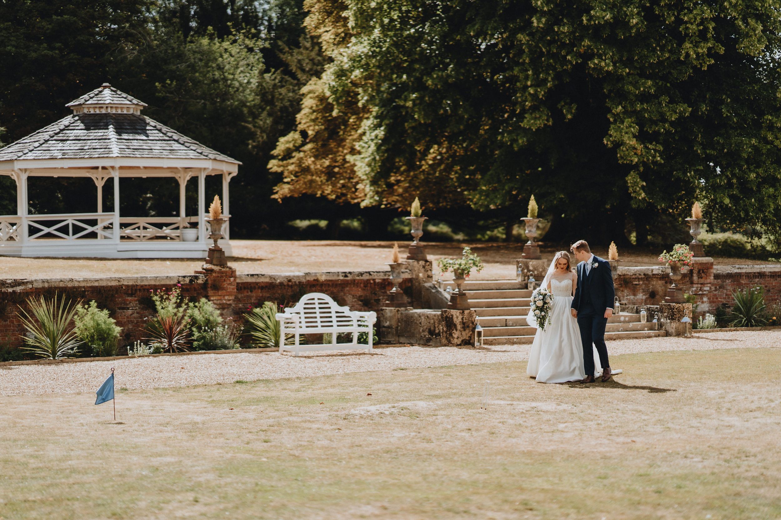 Couple stands on manicured lawn near white wooden gazebo at Holmewood Hall and garden benches during outdoor wedding ceremony.