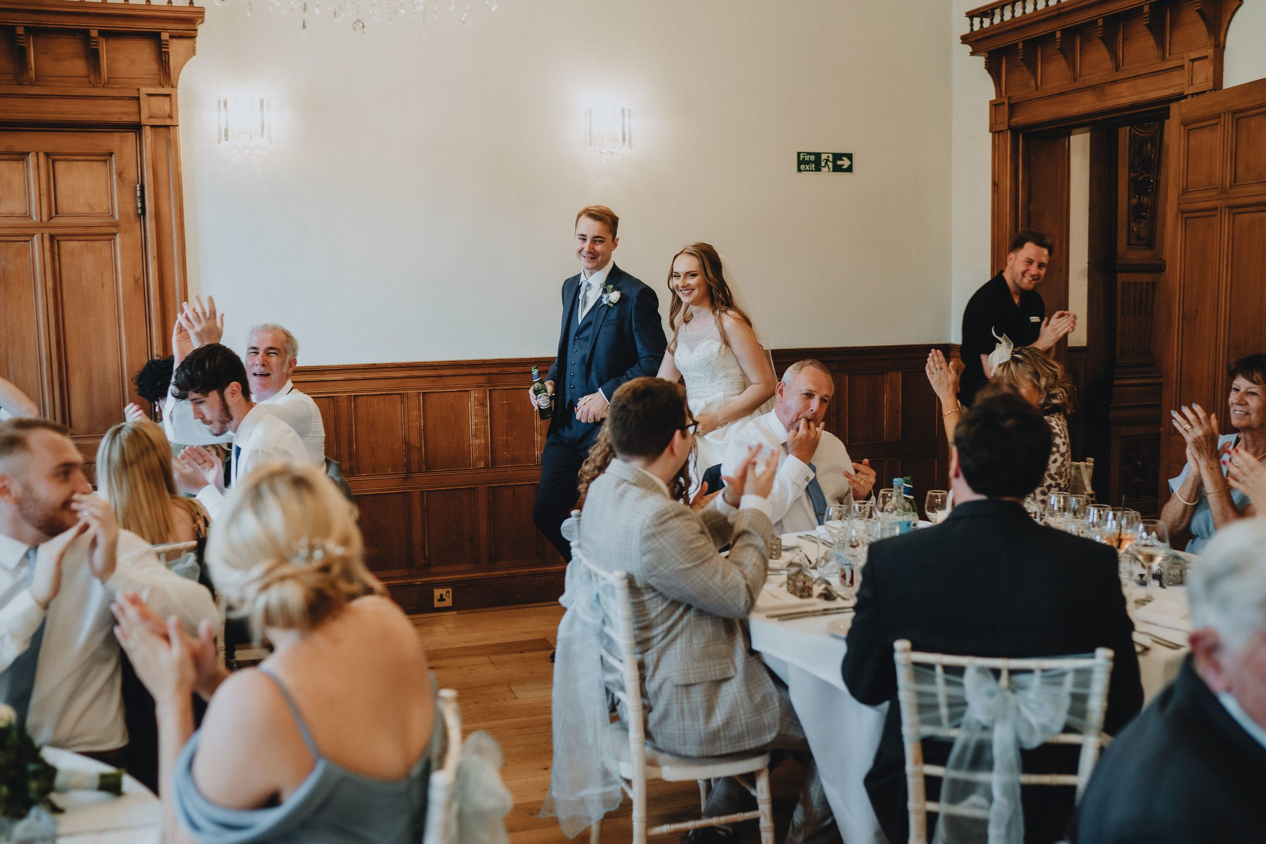A wedding reception celebration in a grand wood-paneled hall with guests seated at round tables for dinner.