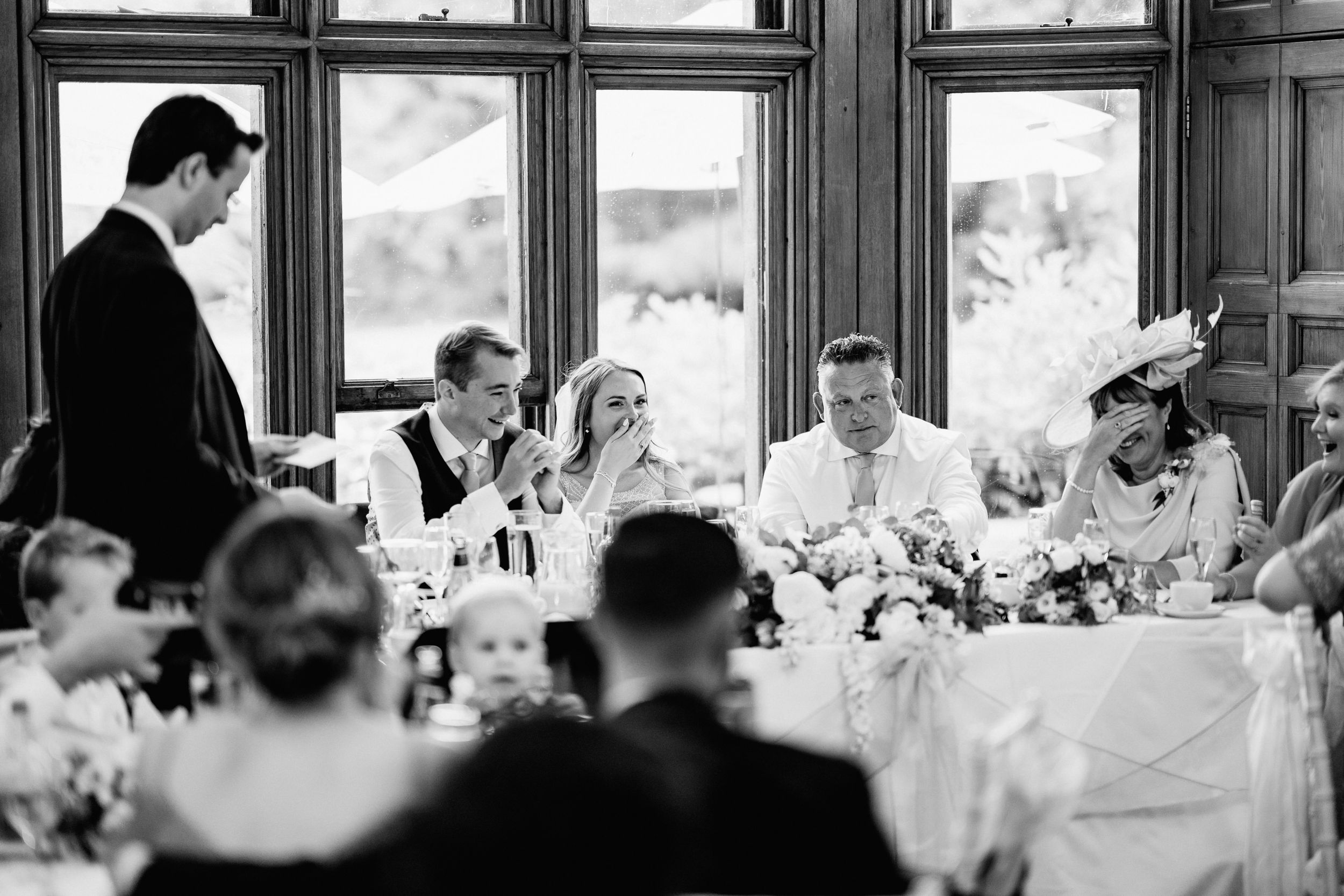 Wedding guests laugh and enjoy themselves during speeches at an elegant reception in a black and white photograph.