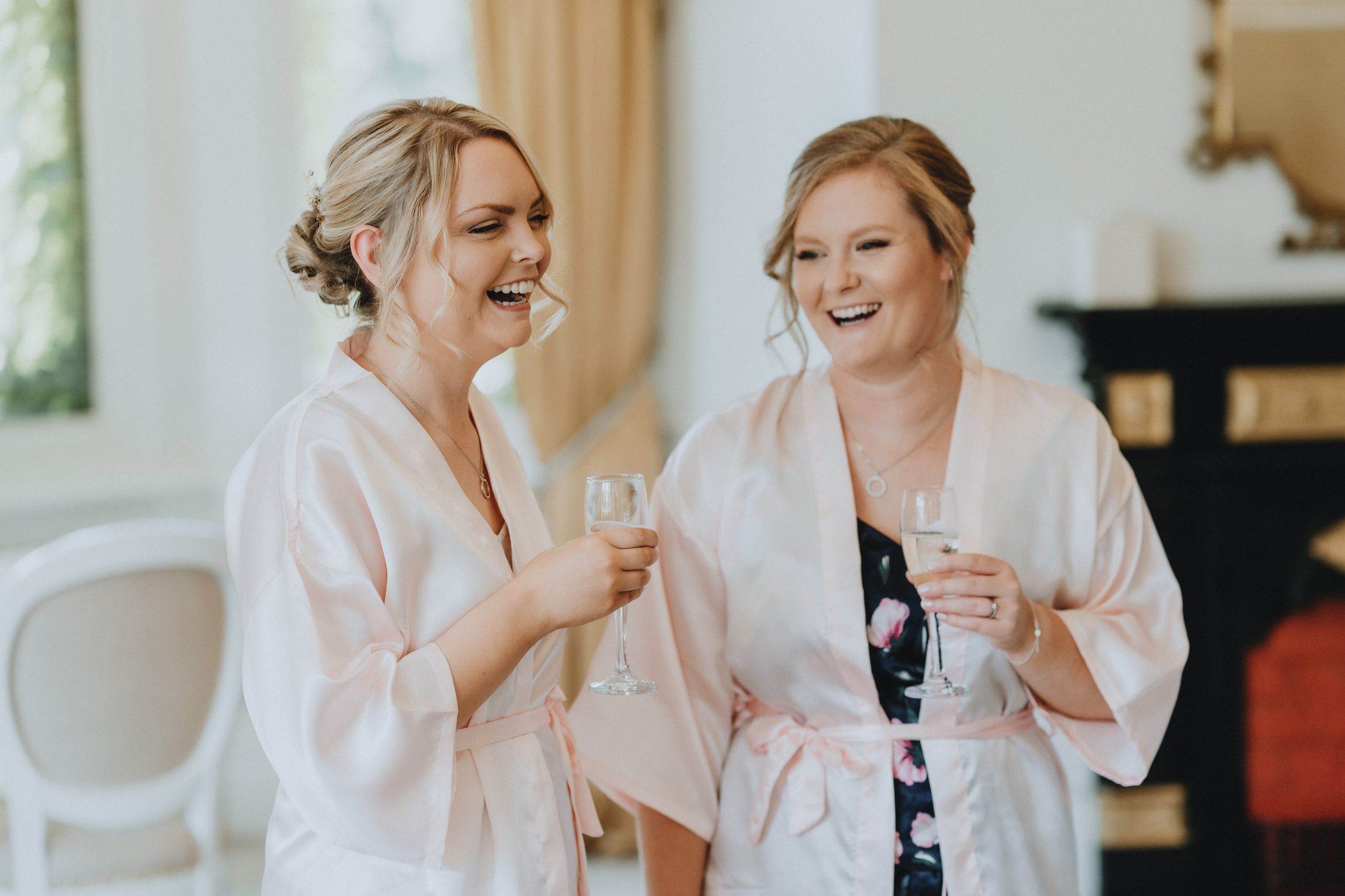 Two women in matching pink robes share a laugh while holding drinks during a wedding preparation moment.