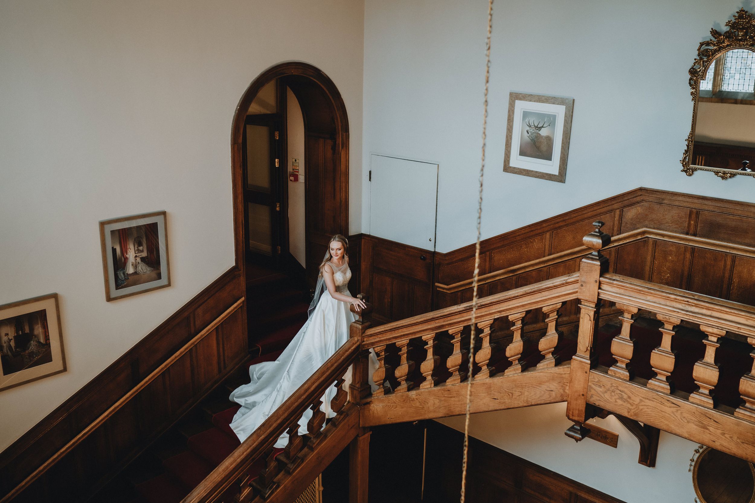 Bride in a flowing white wedding dress stands on a grand wooden staircase at Holmewood Hall, elegant historic building.