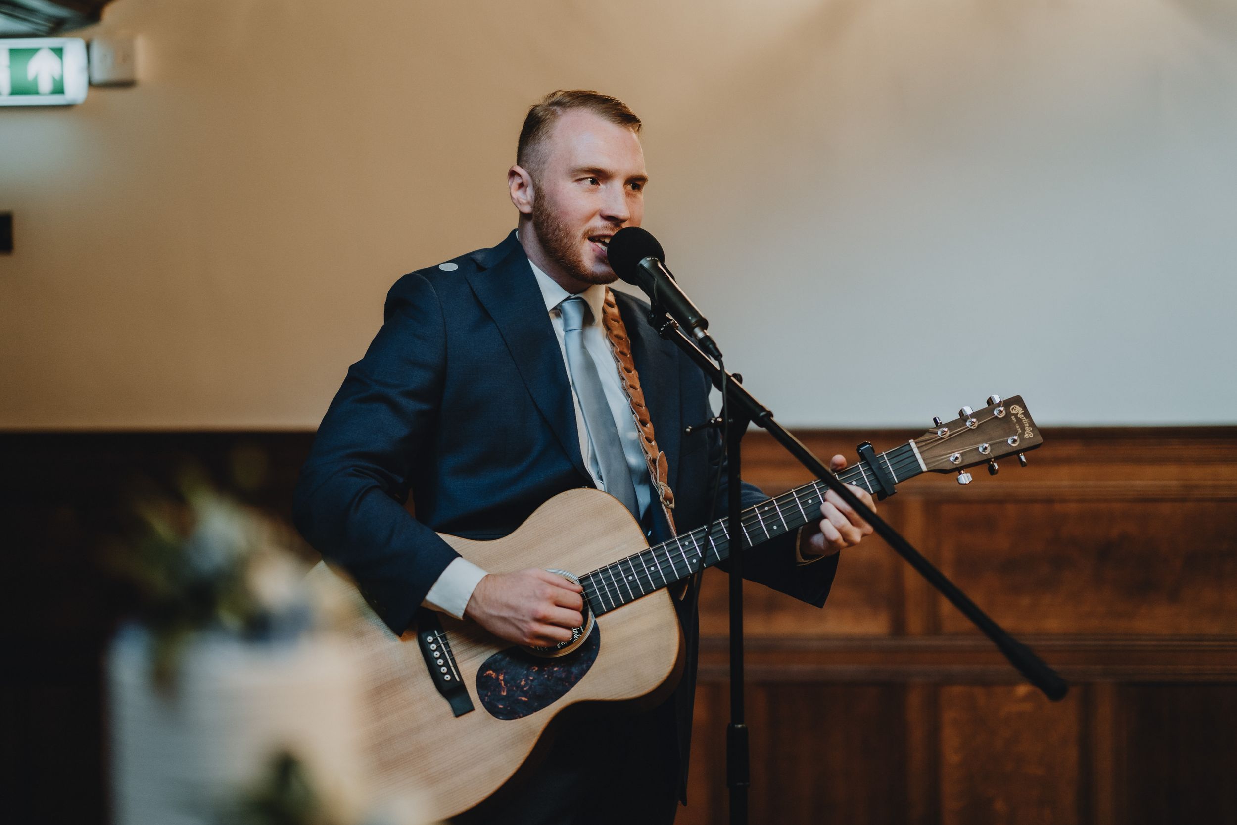 Wedding performer plays acoustic guitar and sings into microphone during indoor reception.
