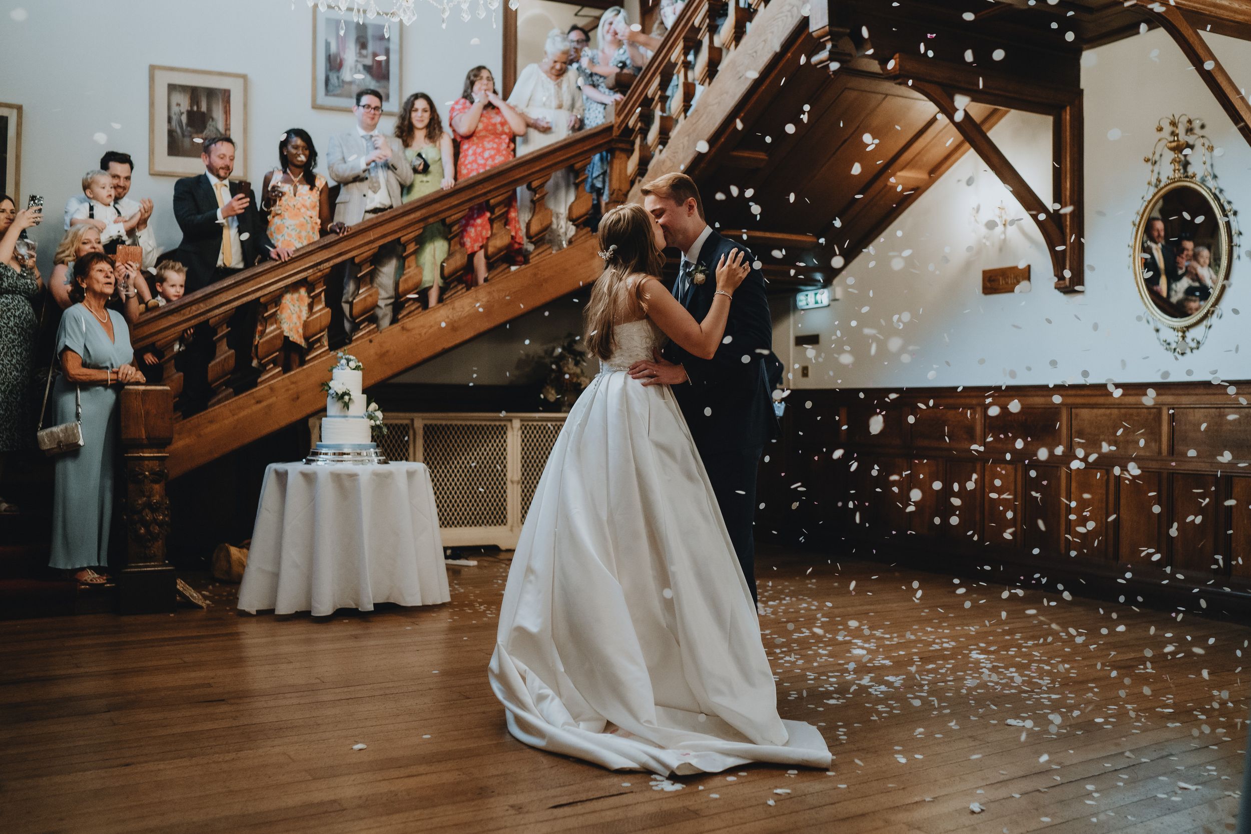 Wedding celebration with guests on wooden staircase watching as confetti falls around flowing white wedding gown in rustic venue.