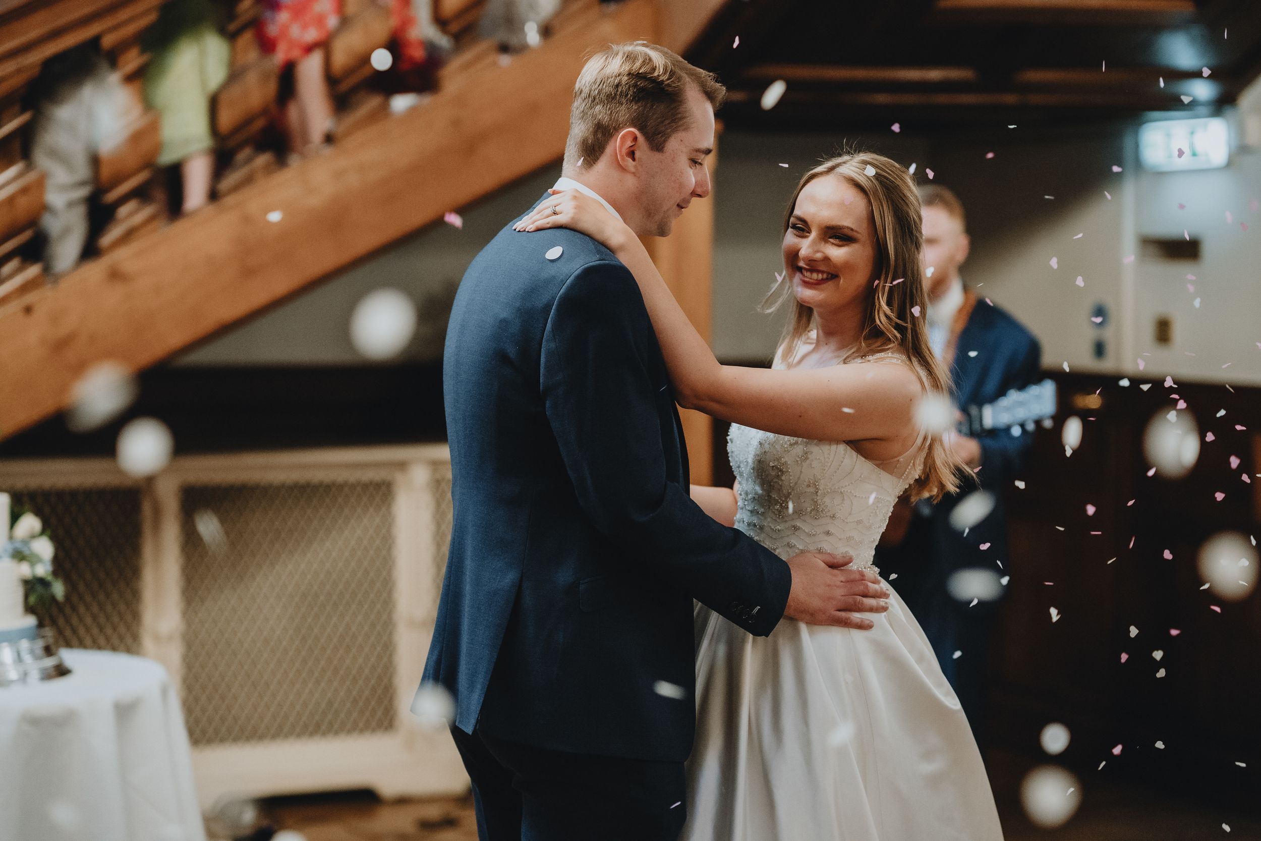 A couple shares their first dance together at a rustic indoor wedding venue with twinkling lights in the background.