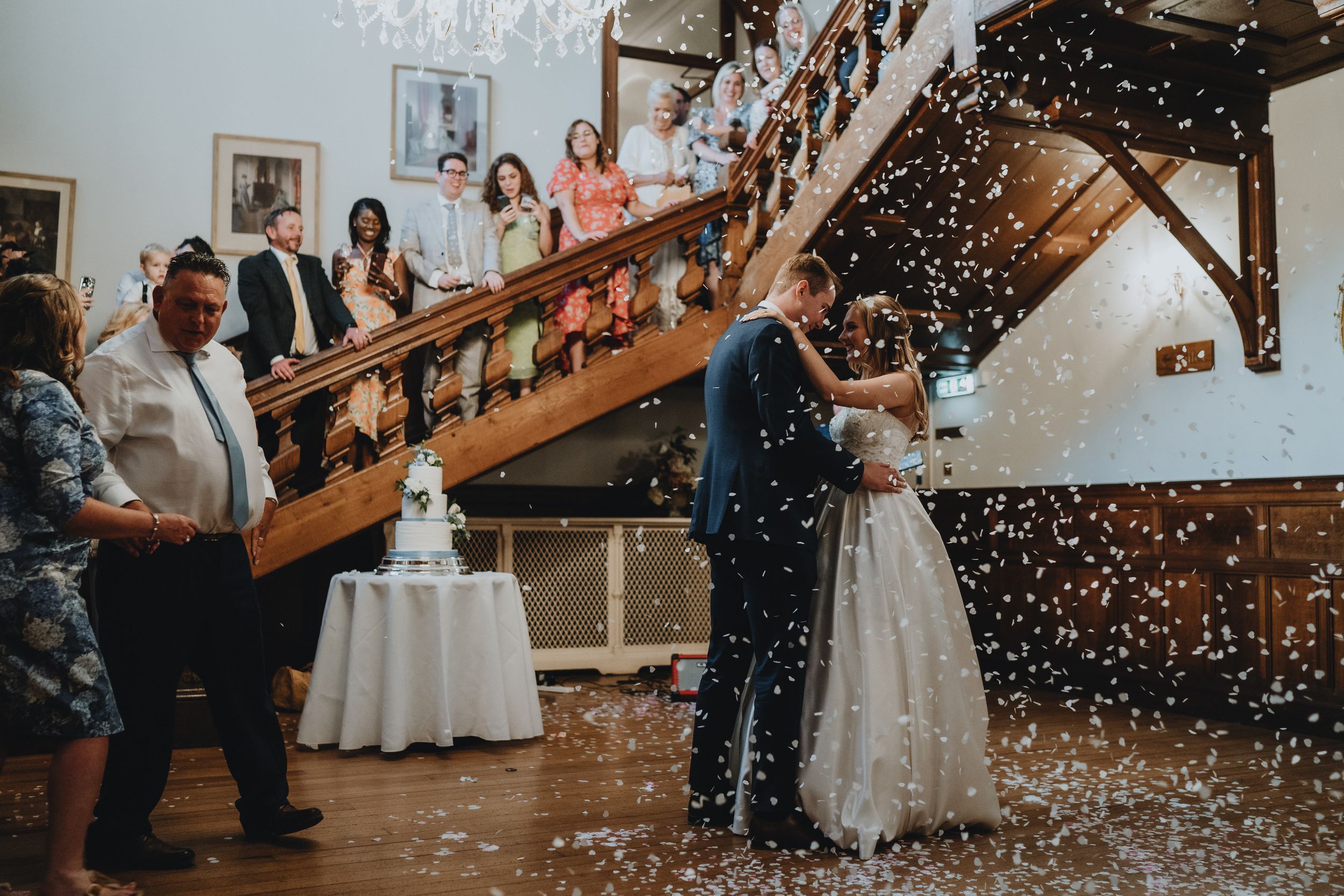 A couple celebrates at Holmewood Hall during their first dance as confetti falls around them in an elegant indoor venue with a staircase.