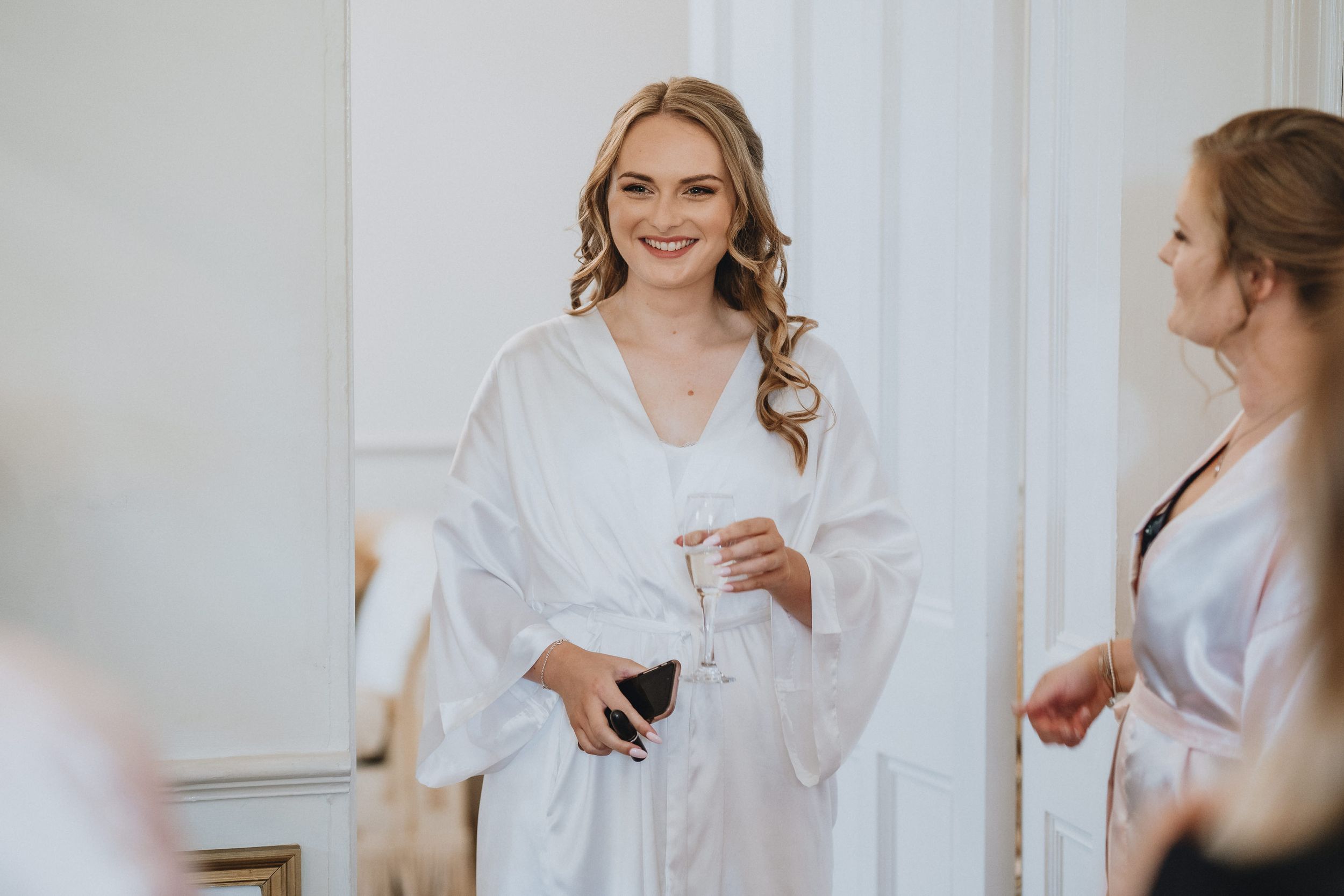 Woman in white silk robe smiles while getting ready in elegant dressing room with mirror.